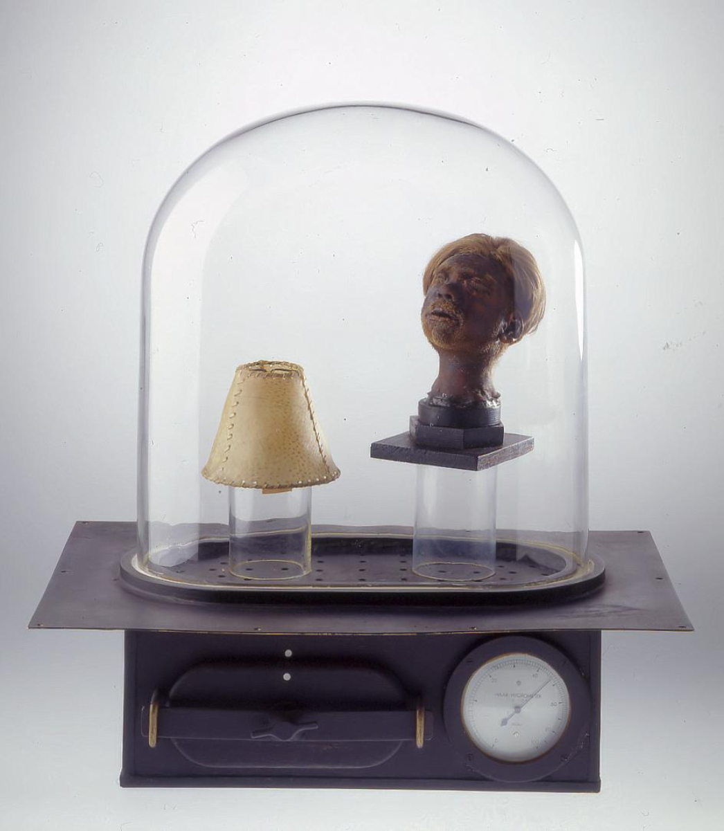 Ein medizinisch-technisches Ausstellungsobjekt unter einer Glasglocke. Unter der Glocke befinden sich zwei makabre Objekte: links ein kleiner Lampenschirm, offenbar aus menschlicher Haut gefertigt, mit sichtbaren Nähten; rechts ein präparierter menschlicher Kopf mit intakter Kopfhaut und Haaren, auf einem Podest montiert. Die Glasglocke steht auf einer Apparatur mit einem Manometer (Druckmesser) und einer Schublade, möglicherweise zur Konservierung oder Präsentation anatomischer Präparate. Das Objekt verweist auf die menschenverachtenden Praktiken im Konzentrationslager Buchenwald und symbolisiert die extreme Entmenschlichung der Opfer.