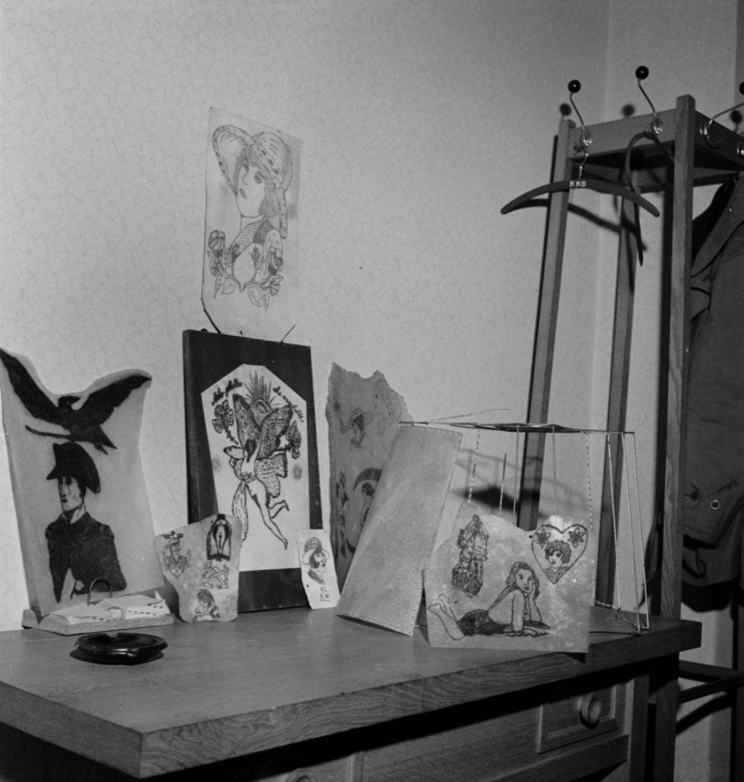 Ein Schwarz-Weiß-Foto zeigt eine Sammlung von Objekten auf einem Holztisch in einem Raum mit Garderobe. Zu sehen sind mehrere Tätowierungen auf scheinbaren Hautstücken, sorgfältig ausgestellt und teils gerahmt oder an eine Konstruktion gehängt. Dargestellt sind unter anderem Porträts, Frauenfiguren und dekorative Motive. Daneben steht ein Lampenschirm, der offenbar ebenfalls aus tätowierter menschlicher Haut gefertigt ist. Das Bild dokumentiert eine makabre Sammlung, die höchstwahrscheinlich aus der NS-Zeit stammt und zur Veranschaulichung nationalsozialistischer Verbrechen an Häftlingen diente.