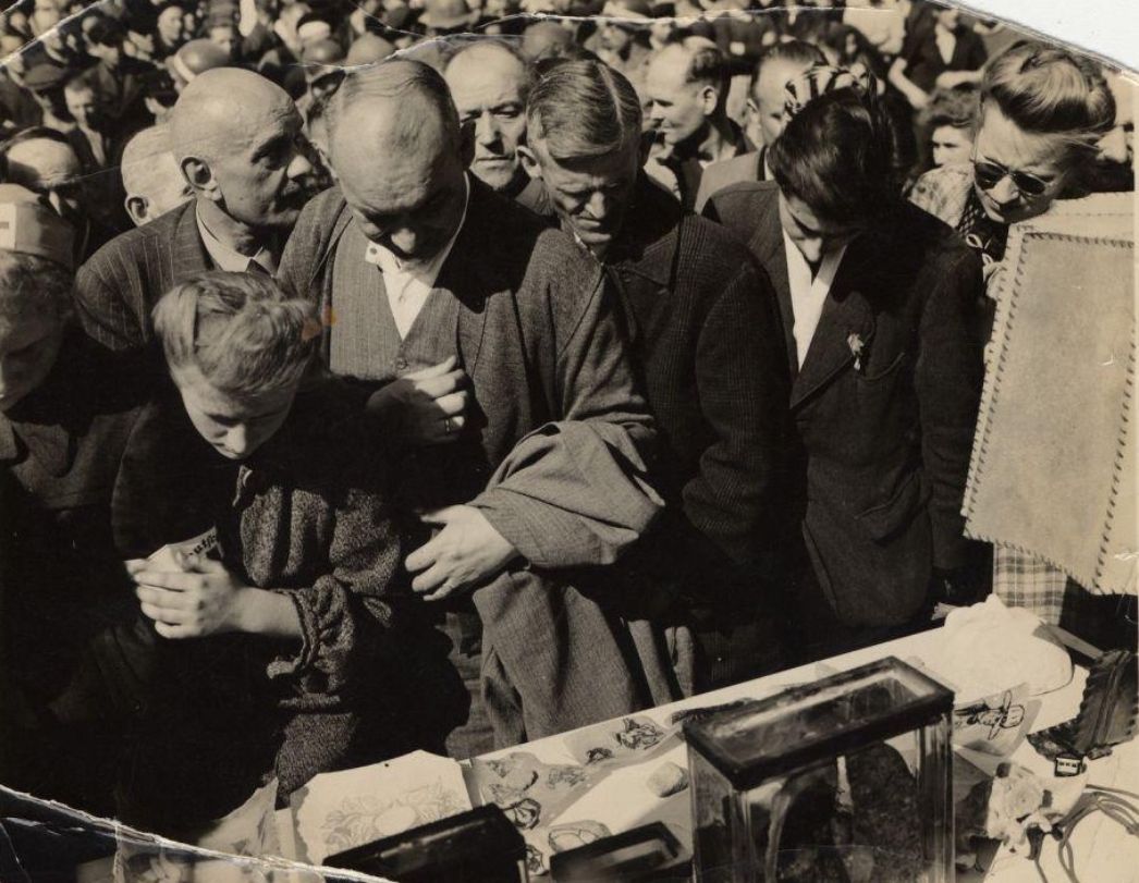 Schwarzweißfotografie einer Menschenmenge, die sich eng um einen Tisch drängt. Im Vordergrund blicken Männer und Frauen mit ernsten, teils erschrockenen Mienen auf ausgestellte Objekte. Auf dem Tisch liegen und stehen anatomische Präparate, darunter ein Glasbehälter mit einem menschlichen Organ, Tätowierungen auf Hautfragmenten sowie weitere makabre Exponate. Rechts im Bildrand ist eine Tischlampe aus Menschenhaut mit erkennbaren Nähten zu sehen. Die Szene spielt sich offenbar nach der Befreiung des Konzentrationslagers Buchenwald ab, als die NS-Verbrechen der Öffentlichkeit zugänglich gemacht wurden.