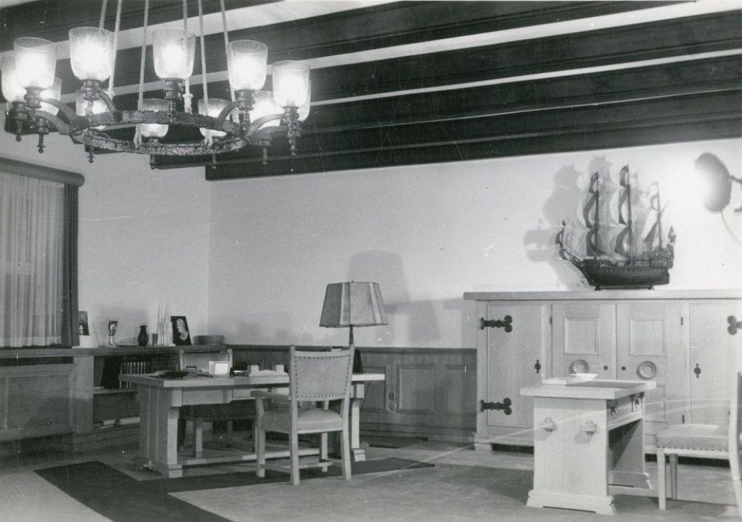 Schwarzweiß-Fotografie eines historischen Bürozimmers mit massivem Holzmobiliar: Im Raum stehen zwei Schreibtische mit Stühlen, eine große Kommode mit einem Modellsegelschiff darauf und eine Stehlampe. An der Decke hängt ein Kronleuchter mit mehreren Glaslampen. Der Raum wirkt geordnet und repräsentativ.