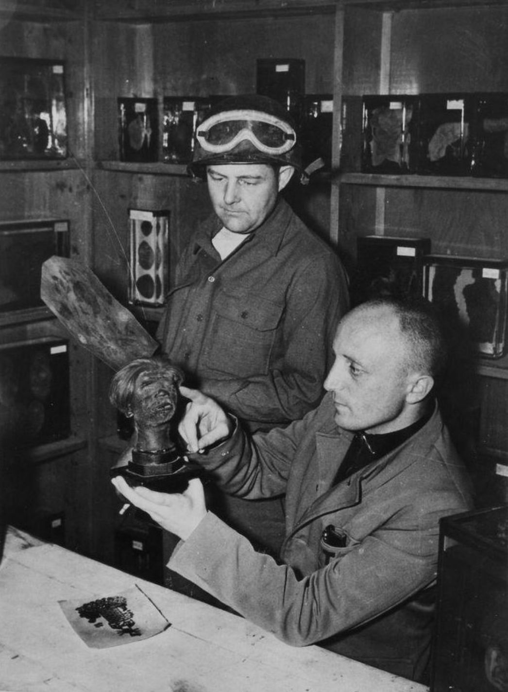 Zwei Männer in Arbeitskleidung, einer mit Schutzbrille auf dem Kopf, befinden sich in einem Raum mit Regalen voller Einweckgläser, die anatomische Präparate enthalten. Der sitzende Mann hält eine kleine, realistisch wirkende Büste mit menschlichen Gesichtszügen in den Händen und zeigt mit dem Finger auf ein Detail, während der stehende Mann aufmerksam zusieht. Auf dem Tisch liegt ein weiteres Objekt, vermutlich Haut mit tätowiertem Motiv.