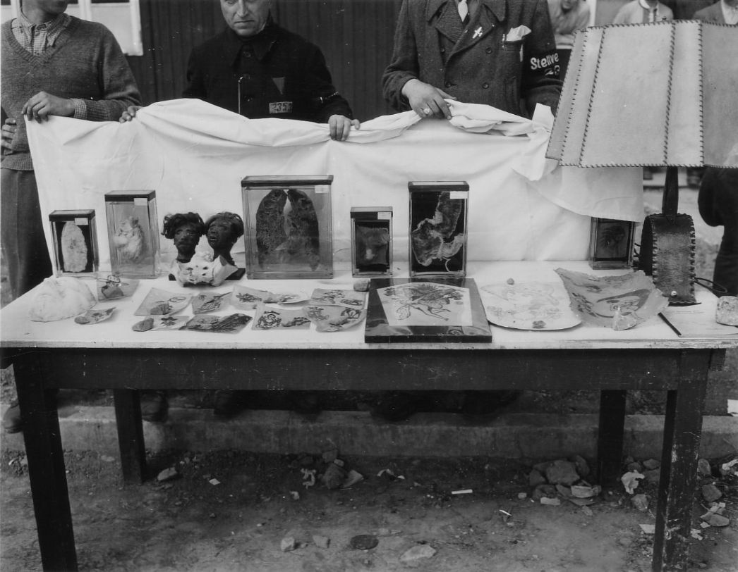 Schwarz-Weiß-Fotografie eines Tisches im Freien, auf dem makabre Exponate präsentiert werden. In Glaskästen befinden sich konservierte menschliche Organe und Körperteile, darunter tätowierte Haut und Schrumpfköpfe. Daneben liegen bemalte Pergamentstücke und eine Lampe mit einem Schirm aus Menschenhaut. Drei Männer, zwei von ihnen mit Häftlingskleidung und sichtbar tätowierten Nummern oder Stoffkennzeichen, halten im Hintergrund ein weißes Tuch als neutrale Präsentationsfläche. Das Bild dokumentiert die Ausstellung von Objekten, die im Konzentrationslager Buchenwald gefunden wurden.