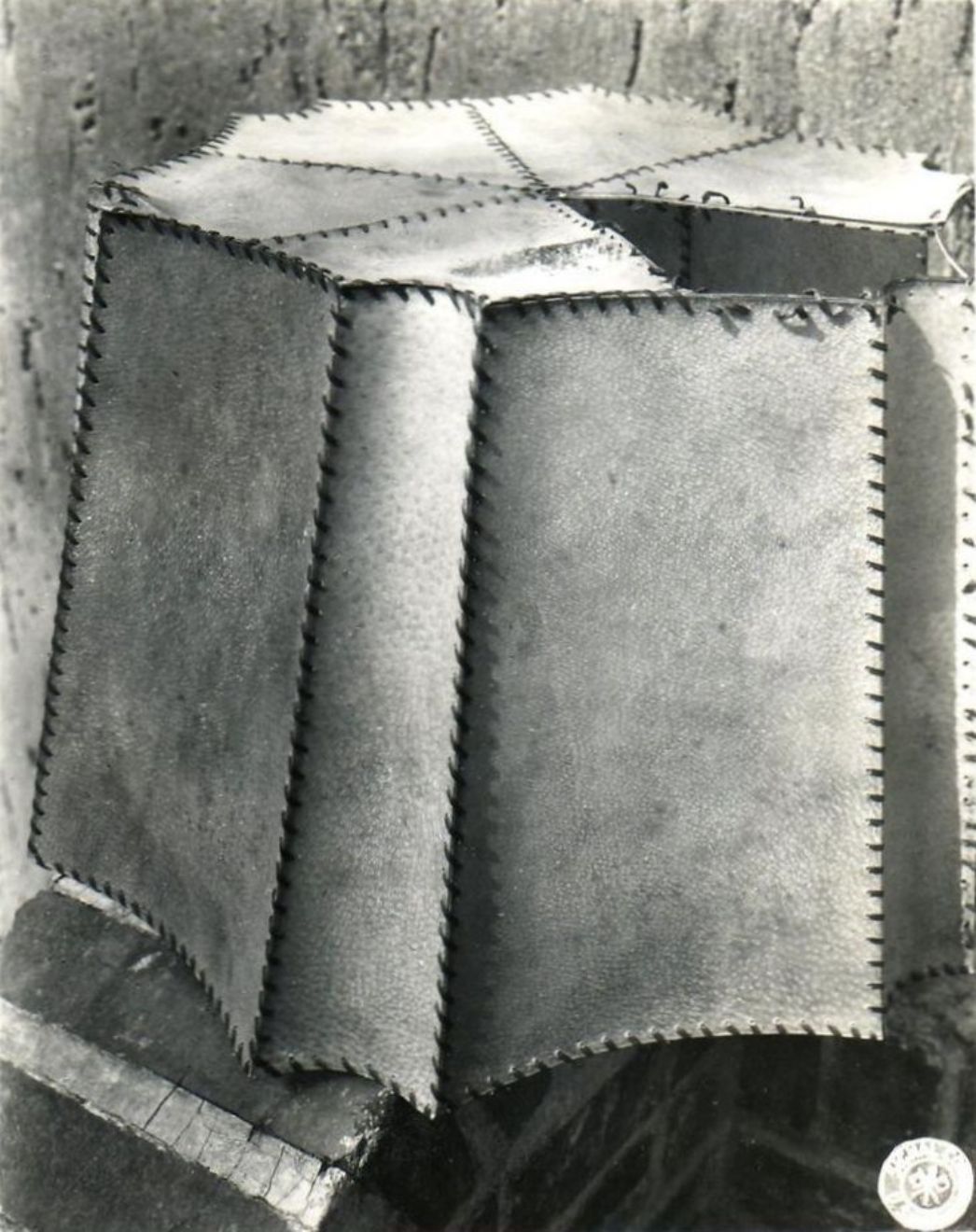 Schwarzweiß-Fotografie eines sechseckigen Lampenschirms, der aus einzelnen rechteckigen Segmenten zusammengesetzt ist. Die Nähte der Segmente sind mit groben Stichen deutlich sichtbar. Der Lampenschirm steht auf einem dunklen Untergrund und wirkt handgefertigt.