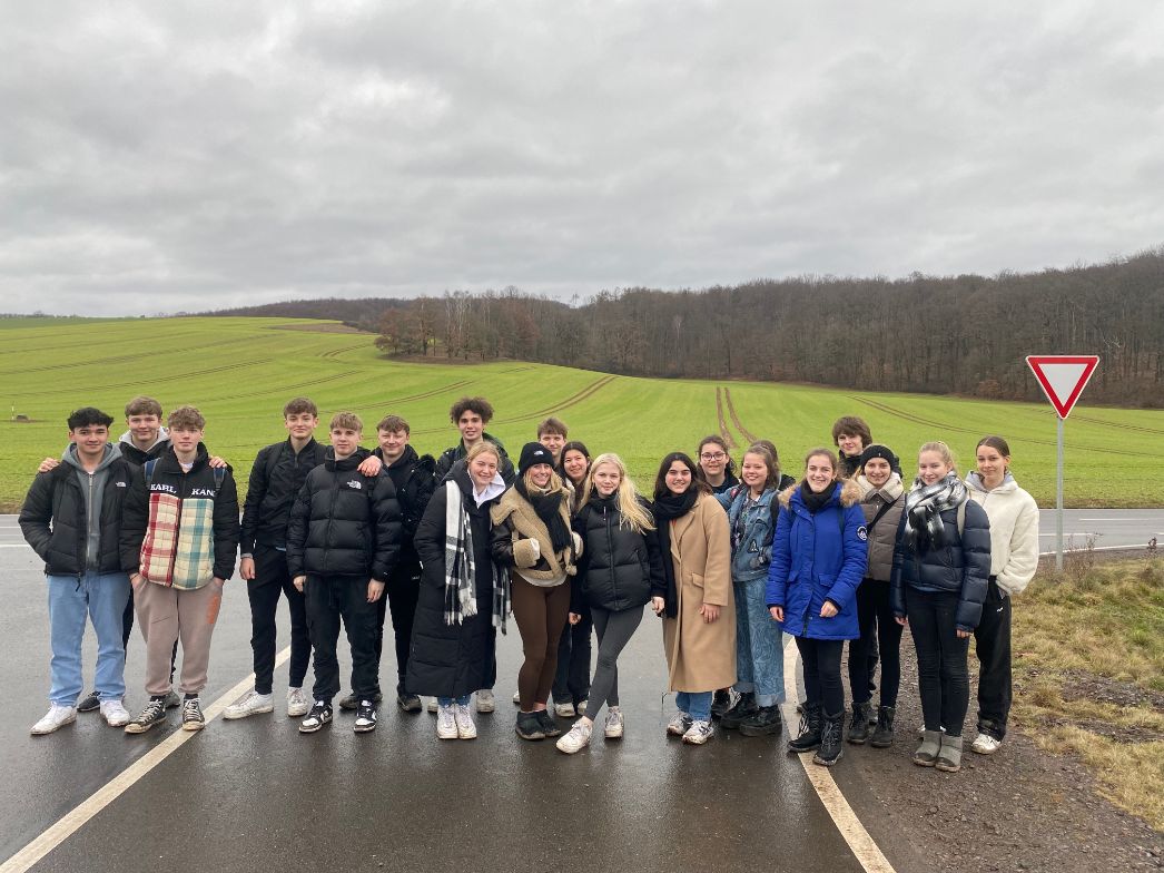 Gruppe von Schülerinnen und Schülern steht auf einer Landstraße vor einem grünen Feld mit Fahrspuren; im Hintergrund Wald unter grauem, bewölktem Himmel, rechts ein Verkehrszeichen (Vorfahrt gewähren).