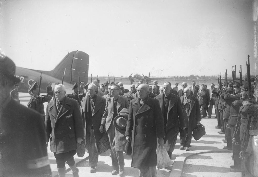 Diese Aufnahme zeigt eine Gruppe ehemaliger Häftlinge, die gerade an einem Flughafen eintreffen oder abreisen – vermutlich im Zuge ihrer Repatriierung nach der Befreiung aus einem Konzentrationslager oder einem Kriegsgefangenenlager. Im Hintergrund ist ein Transportflugzeug (vermutlich eine Douglas C-47 „Skytrain“) zu sehen, ein typisches Modell der Alliierten zur Zeit des Zweiten Weltkriegs. Die Männer im Vordergrund wirken älter, viele tragen einfache Mäntel und halten persönliche Gegenstände in der Hand – Beutel, Taschen oder Mützen. Einige tragen noch Kennzeichnungen auf der Kleidung, möglicherweise Häftlingsnummern oder Abzeichen. Beidseitig werden sie von Soldaten empfangen, die in einer Ehrenformation mit aufgepflanztem Bajonett Spalier stehen – ein Zeichen des Respekts und der Anerkennung für das durchlittene Leid dieser Männer. Diese Szene steht symbolisch für die Rückkehr in die Freiheit und die beginnende Aufarbeitung der Verbrechen des NS-Regimes. Der Moment ist feierlich, würdevoll, und drückt gleichzeitig die Erleichterung und den Neuanfang aus.