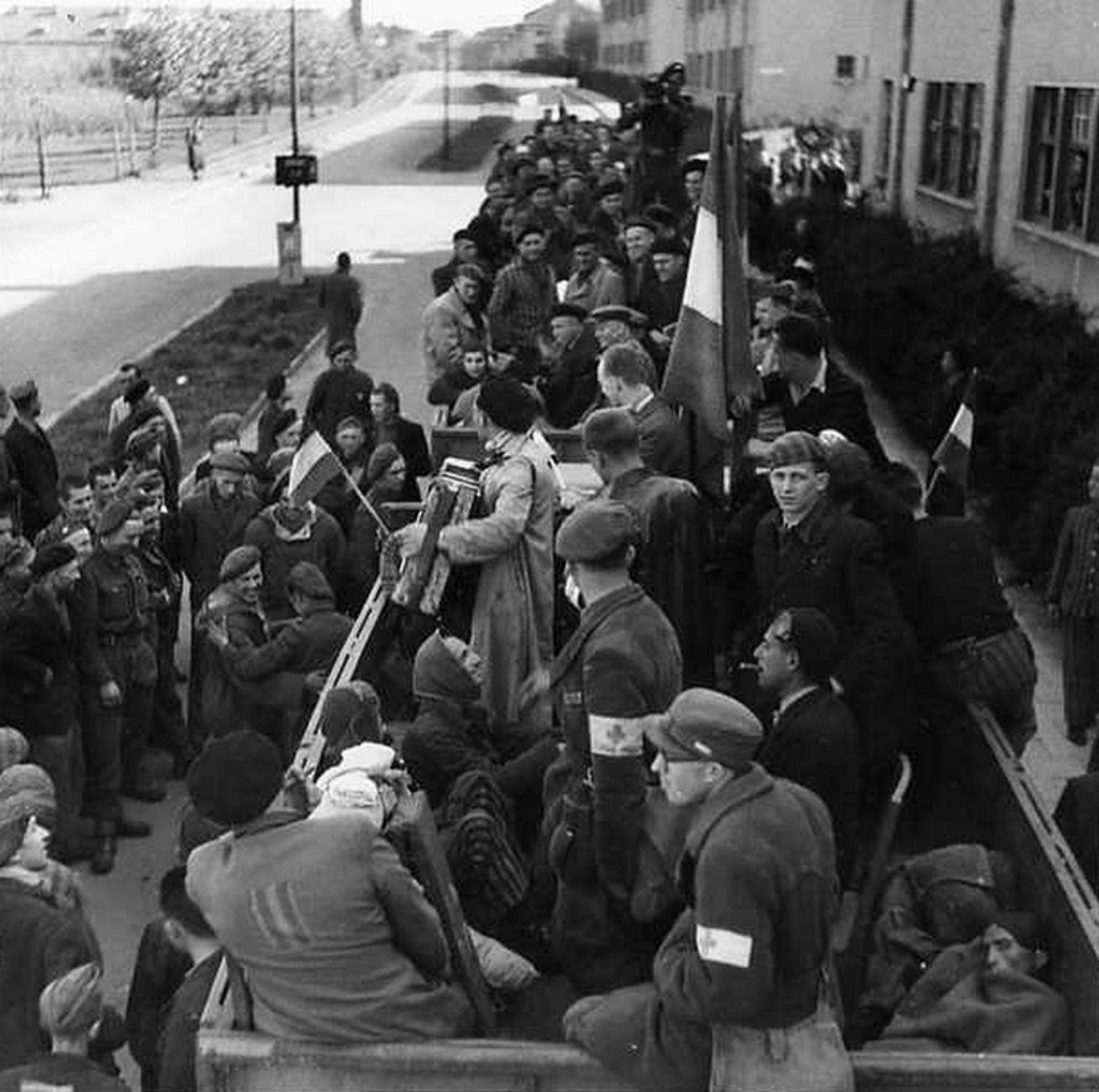 Dieses Bild zeigt eine Szene der Evakuierung oder Rückführung befreiter KZ-Häftlinge – wahrscheinlich aus dem Konzentrationslager Dachau oder einem seiner Außenlager. Die Aufnahme stammt sehr wahrscheinlich aus dem Frühjahr 1945, kurz nach der Befreiung. Zu sehen sind zahlreiche Menschen, viele in gestreifter Lagerkleidung oder einfacher ziviler Kleidung. Sie sitzen oder stehen auf einem großen offenen Lastwagen. Deutlich sichtbar sind auch mehrere französische Flaggen sowie Männer mit weißen Armbinden – ein typisches Erkennungsmerkmal für medizinisches Personal, Mitglieder des Roten Kreuzes oder der Résistance. In der Bildmitte spielt jemand auf einem Akkordeon – ein ergreifender Moment, der trotz der entsetzlichen Vorgeschichte einen Hauch von Menschlichkeit, Erleichterung oder sogar Hoffnung vermittelt. Viele schauen in Richtung der Kamera oder begrüßen sich gegenseitig. Am Bildrand stehen alliierte Soldaten, vermutlich amerikanische Befreier, in Uniform. Diese Szene dokumentiert eindrucksvoll den Moment der Rückkehr in die Freiheit. Die Aufnahme gehört zur Kategorie der humanitären Nachkriegsfotografie, die nicht nur das Grauen, sondern auch den Anfang des Wiederaufbaus und der Rückkehr zur Würde zeigt.