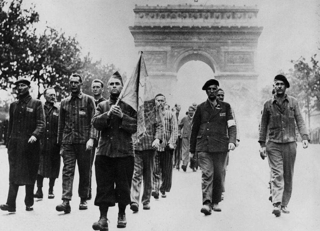 Diese historische Fotografie zeigt eine Gruppe befreiter KZ-Häftlinge, die in Paris auf den Champs-Élysées marschieren, im Hintergrund deutlich erkennbar der Arc de Triomphe. Das Bild ist ein starkes Symbol der Befreiung und des Sieges über den Nationalsozialismus. Viele der Männer tragen noch die gestreifte Häftlingskleidung der Konzentrationslager, einige haben bereits andere Kleidungsstücke erhalten. Der Mann in der Mitte vorne trägt eine Fahne, vermutlich die französische Trikolore – ein Akt des Wiedergewinns von Würde, Freiheit und nationaler Identität. Begleitet werden sie von Angehörigen der Résistance oder der Alliierten, zu erkennen an den Armbinden und Baretten.