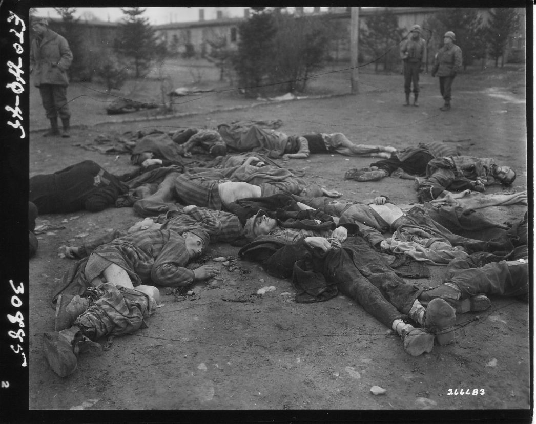 Dieses erschütternde Bild zeigt die Leichen ermordeter Häftlinge eines Konzentrationslagers, vermutlich kurz nach der Befreiung durch alliierte Truppen. Viele der Toten tragen gestreifte Häftlingskleidung, einige sind nur notdürftig bedeckt oder liegen in schmerzverzerrter Haltung am Boden. Im Hintergrund sind amerikanische Soldaten zu sehen, die das Lager gesichert oder gerade entdeckt haben.