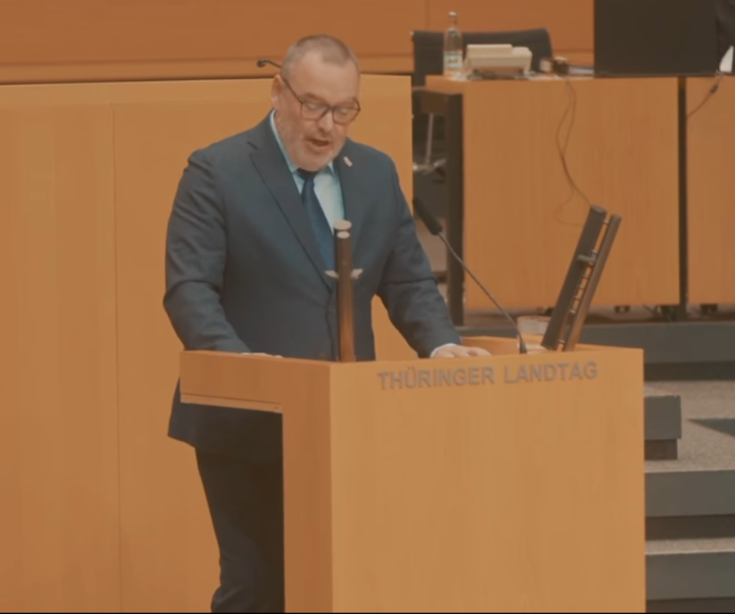 Auf dem Bild ist ein Mann zu sehen, der an einem Rednerpult mit der Aufschrift „THÜRINGER LANDTAG“ spricht. Er trägt einen dunkelblauen Anzug mit einem türkisfarbenen Hemd und passender Krawatte sowie eine Brille. Aufgrund der visuellen Ähnlichkeit mit dem Facebook-Beitrag zuvor handelt es sich sehr wahrscheinlich um Sascha Schlösser, einen Landtagsabgeordneten der AfD Thüringen. Das Setting zeigt eine typische Szene aus einer Parlamentssitzung im Landtag von Thüringen, erkennbar an der Inneneinrichtung und dem Aufbau des Plenarsaals.