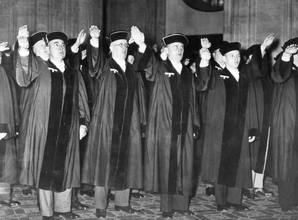 Schwarz-weiß-Fotografie einer Gruppe von Männern in richterlicher Amtstracht, die während des Nationalsozialismus den Hitlergruß zeigen. Die Männer stehen in einer Reihe in einem prunkvollen Innenraum mit verziertem Boden, tragen Roben und Baretts.