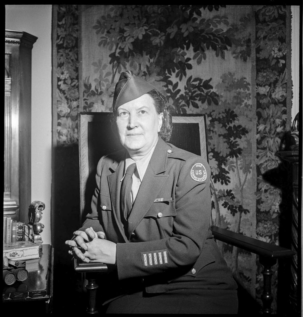 Das Schwarzweißporträt zeigt eine Frau mittleren Alters in Uniform der US-amerikanischen Women's Army Corps (WAC). Sie sitzt auf einem Stuhl mit gefalteten Händen und blickt ruhig in die Kamera. Auf ihrem linken Arm ist ein rundes Abzeichen mit der Aufschrift „U.S. War Correspondent“ zu erkennen. Der Hintergrund zeigt einen dekorativen Wandteppich mit Blattmotiven.