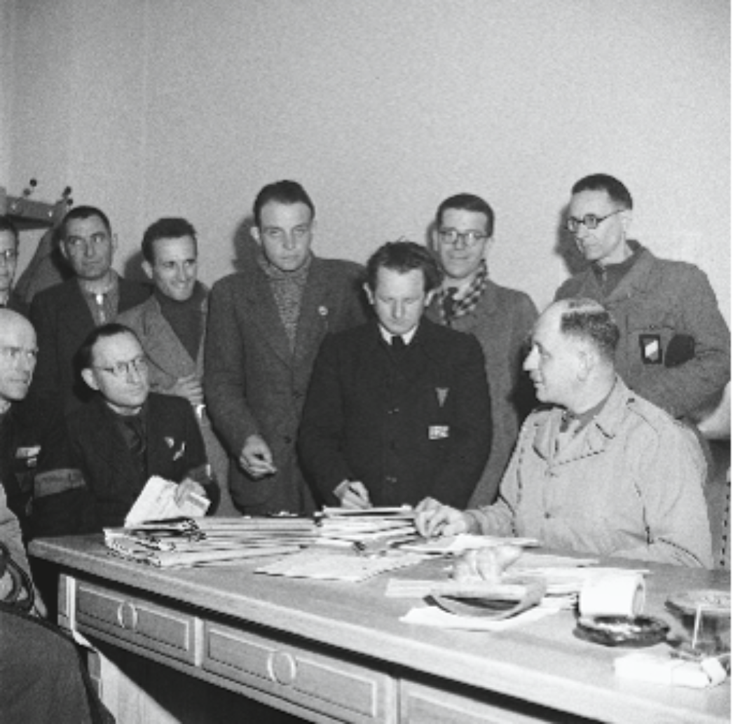 Das Schwarzweißbild zeigt eine Gruppe von Männern in einem schlicht eingerichteten Raum. Ein amerikanischer Offizier sitzt an einem Schreibtisch mit vielen Papieren, während mehrere Zivilisten oder ehemalige Häftlinge um ihn herumstehen. Einige tragen Abzeichen oder Dreiecke an ihrer Kleidung – Hinweise auf ihre frühere Internierung.