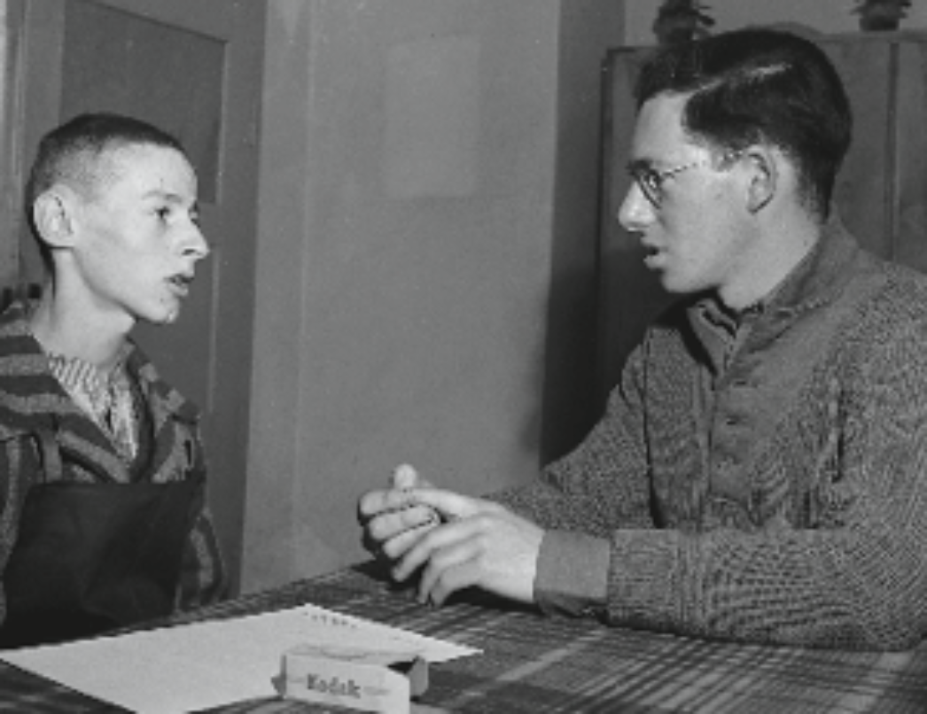 Dieses Foto scheint ein Gespräch oder Interview zwischen zwei Personen in einem historischen Kontext darzustellen. Die Person auf der linken Seite sieht sehr jung aus und trägt gestreifte Kleidung, die an die Häftlingsuniformen aus Konzentrationslagern während des Zweiten Weltkriegs erinnert. Die Person auf der rechten Seite scheint ein Interview zu führen oder Notizen zu machen – möglicherweise ein Journalist, Forscher oder Helfer. Auf dem Tisch liegt eine Schachtel mit dem Schriftzug „Kodak“, was auf fotografische Ausrüstung oder Materialien hinweist – ein weiteres Indiz dafür, dass hier etwas dokumentiert wurde.