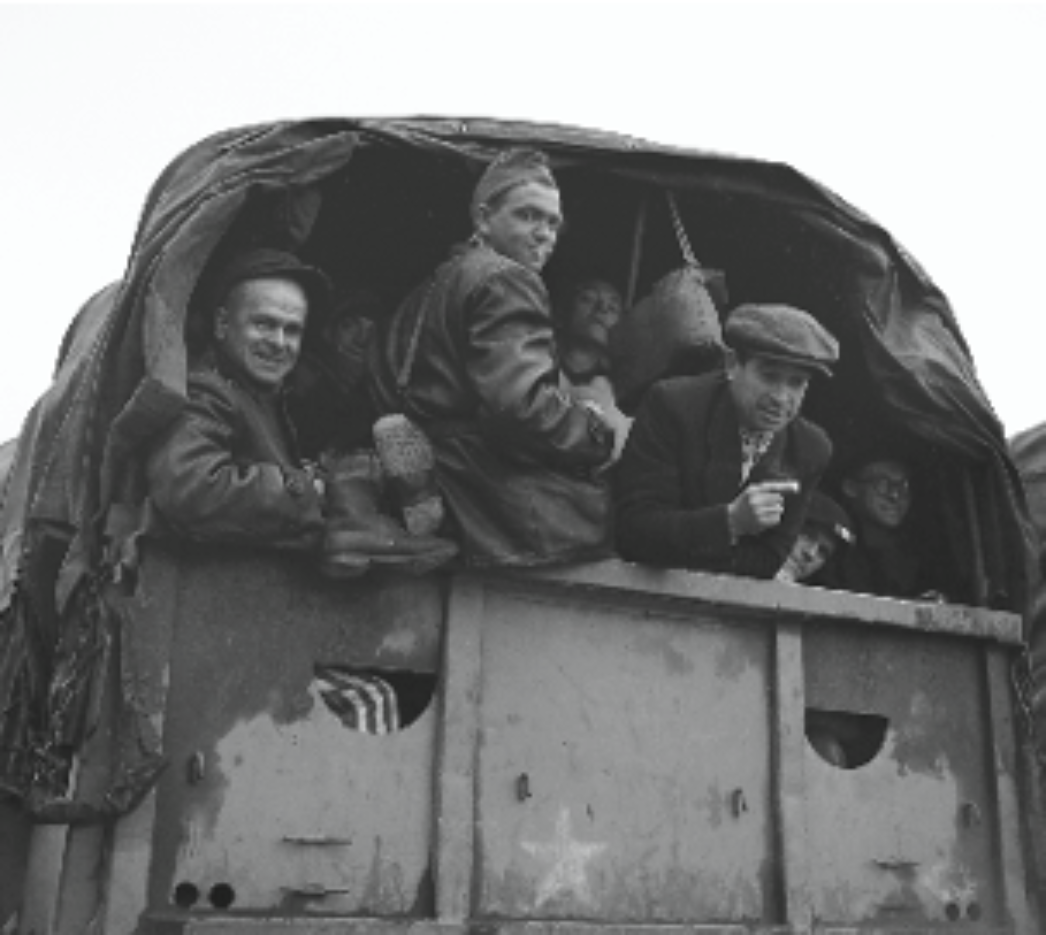 Dieses Bild zeigt eine Gruppe von Männern auf der Ladefläche eines offenen Lastwagens. Die Szene wirkt historisch und erinnert stark an die Zeit des Zweiten Weltkriegs oder kurz danach. Einige Männer tragen Kleidung, die an ehemalige KZ-Häftlinge erinnert (z. B. gestreifte Stoffe, sichtbar am unteren Rand). Andere tragen zivile Kleidung oder einfache Arbeitskleidung. Die Stimmung wirkt trotz der offensichtlichen Härte der Situation relativ positiv – einige Männer lächeln oder schauen neugierig aus dem Fahrzeug. Es könnte sich um Überlebende handeln, die gerade befreit wurden und auf dem Weg aus einem Konzentrationslager oder zu einem Auffanglager sind. Die Kombination mit dem ersten Bild legt nahe, dass es sich hier um eine Serie von Fotos handelt, die die Befreiung und Dokumentation von KZ-Häftlingen durch Alliierte oder Hilfsorganisationen zeigt.