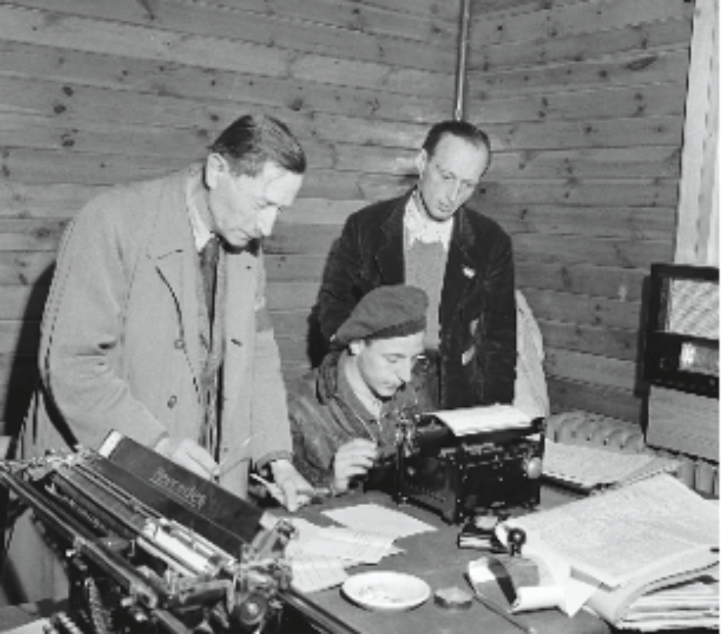 Dieses Bild zeigt drei Männer in einem Büro oder Verwaltungsraum, vermutlich in einer provisorischen Unterkunft oder einem Lager, wie es nach dem Zweiten Weltkrieg oft bei der Verwaltung von Displaced Persons (DPs) oder befreiten KZ-Häftlingen der Fall war. Ein Mann sitzt an einer Schreibmaschine, während die beiden anderen Männer stehend über seine Schulter schauen und offenbar Dokumente begutachten oder besprechen. Die Schreibmaschine im Vordergrund trägt die Aufschrift „Remington“, ein bekannter Hersteller. Die Holzverkleidung des Raumes deutet auf eine einfache, temporäre Einrichtung hin – möglicherweise eine Baracke in einem ehemaligen Lager oder einer Hilfseinrichtung.