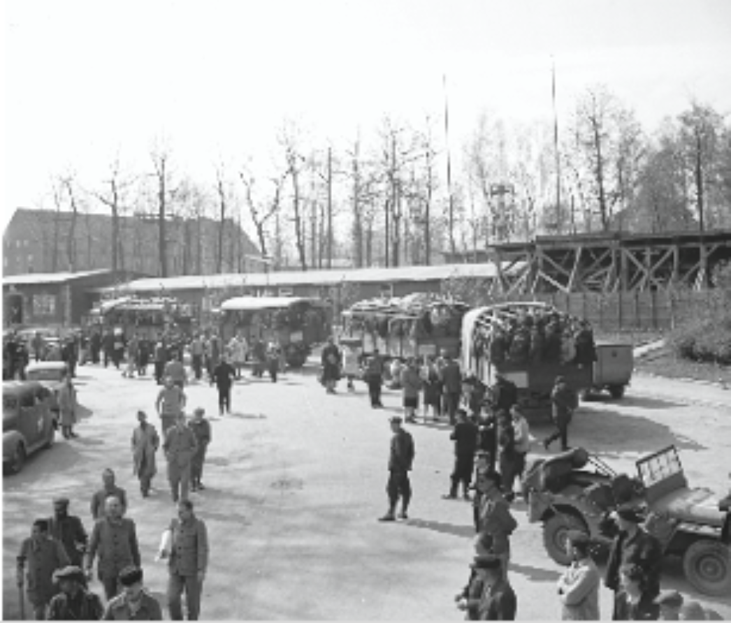 Dieses Bild zeigt eine weiträumige Szene auf einem offenen Platz mit zahlreichen Menschen, Fahrzeugen und Lastwagen. Besonders auffällig ist der Lastwagen rechts, der voll mit Personen beladen ist – vermutlich ehemalige Häftlinge oder Displaced Persons, wie man sie nach der Befreiung der Konzentrationslager nannte. Die Umgebung und der Baustil der Gebäude sowie der improvisierte Holzsteg im Hintergrund deuten darauf hin, dass es sich um ein ehemaliges Lager handelt – möglicherweise ein Konzentrationslager oder ein Auffanglager für Überlebende. Im Vordergrund sind alliierte Soldaten und Zivilisten zu sehen, darunter auch Kinder und Frauen. Die Situation wirkt geordnet, aber geschäftig – vermutlich handelt es sich um eine Evakuierung, Umsiedlung oder Registrierung nach der Befreiung eines Lagers.