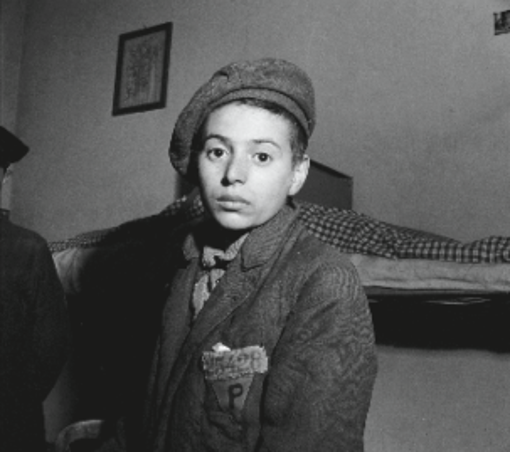 Dieses Foto zeigt einen jungen Überlebenden, vermutlich aus einem Konzentrationslager, kurz nach der Befreiung. Die Kleidung ist abgetragen und notdürftig zusammengesetzt, was typisch ist für KZ-Häftlinge. Auf seiner Jacke ist ein Stoffabzeichen mit einem „P“ zu erkennen – dieses Zeichen wurde im NS-Regime häufig verwendet, um polnische Zwangsarbeiter oder Häftlinge zu kennzeichnen („P“ für „Pole“). Der Blick des Jungen ist ernst und ruhig, was häufig auf die tiefgreifenden Traumata hinweist, die viele junge Überlebende erlitten haben. Im Hintergrund sieht man ein Bett mit kariertem Bettzeug, was auf eine provisorische Unterkunft hinweist – eventuell ein Auffanglager oder eine Baracke nach der Befreiung.