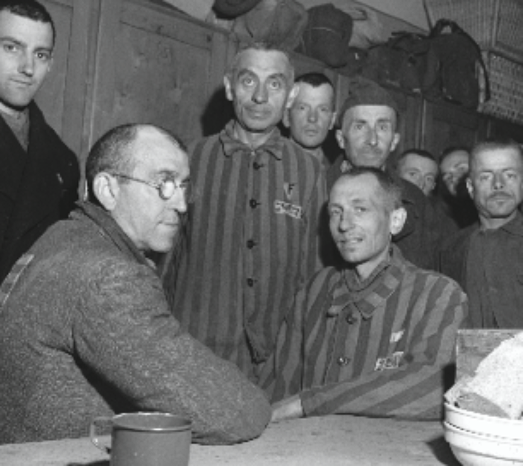 Dieses Bild zeigt eine Gruppe von Männern, von denen mehrere die typische gestreifte Häftlingskleidung aus Konzentrationslagern tragen. Auf den Jacken sind Häftlingsnummern und Abzeichen zu erkennen, die oft zur Kennzeichnung der Haftgründe dienten (z. B. politische Gefangene, Juden, Homosexuelle, Zeugen Jehovas, etc.). Die Männer wirken erschöpft, aber aufmerksam, einige schauen direkt in die Kamera. Im Vordergrund sitzt ein Mann mit Brille an einem Tisch, neben ihm ein Blechnapf oder eine Tasse. Im Hintergrund erkennt man Regale oder persönliche Gegenstände über Spinden – ein Hinweis darauf, dass dieses Foto in einer Unterkunftsbaracke aufgenommen wurde. Die Gesichter und Körper der Männer sind dünn, was auf lange Haft und Mangelernährung hindeutet. Trotzdem scheinen sie sich in einem Moment relativer Ruhe zu befinden, möglicherweise nach der Befreiung.