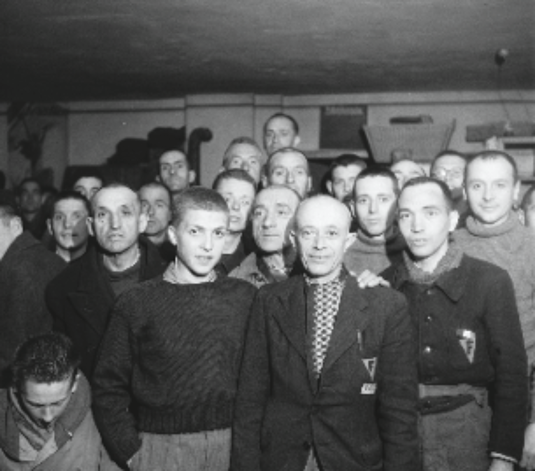 Dieses Bild zeigt eine größere Gruppe von Männern, dicht gedrängt in einem Innenraum. Einige tragen deutlich erkennbare Elemente der Häftlingskleidung aus Konzentrationslagern, z. B. gestreifte Hemden oder Jacken mit aufgenähten Abzeichen. Andere scheinen zivile oder improvisierte Kleidung zu tragen. Besonders auffällig ist der Mann in der Mitte mit Brille und einem Dreieck auf seiner Jacke – vermutlich ein Häftlingsabzeichen, das auf seine Haftkategorie hinweist. Die Gesichter zeigen eine Mischung aus Erschöpfung, Ernsthaftigkeit und vorsichtiger Aufmerksamkeit. Einige blicken direkt in die Kamera, andere zur Seite. Die Szene spielt sich wahrscheinlich in einem Unterkunftsraum ab.
