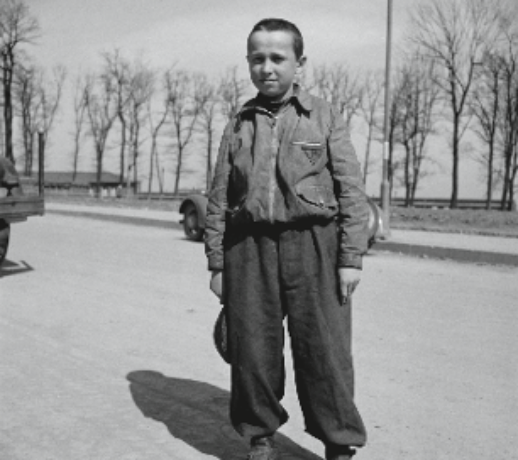 Dieses Bild zeigt einen jungen Jungen, der allein auf einer Straße steht. Er trägt Kleidung, die typisch für ehemalige KZ-Häftlinge ist: ein gestreiftes Hemd und eine Jacke mit einem erkennbaren Häftlingsabzeichen (ein aufgenähtes Dreieck auf der Brust). Die Hose wirkt zu groß, was darauf hindeutet, dass sie notdürftig zur Verfügung gestellt wurde. Im Hintergrund ist eine karge Landschaft mit kahlen Bäumen zu sehen, was auf eine Aufnahme im Frühjahr oder Spätwinter hindeuten könnte – möglicherweise unmittelbar nach der Befreiung eines Lagers. Der Gesichtsausdruck des Jungen ist ruhig, vielleicht erschöpft oder abwartend. Die Szene wirkt isoliert und macht die Verletzlichkeit und Jugend des Jungen besonders deutlich. Die Kleidung, das Abzeichen und die Umgebung lassen darauf schließen, dass er ein Überlebender eines Konzentrationslagers ist.
