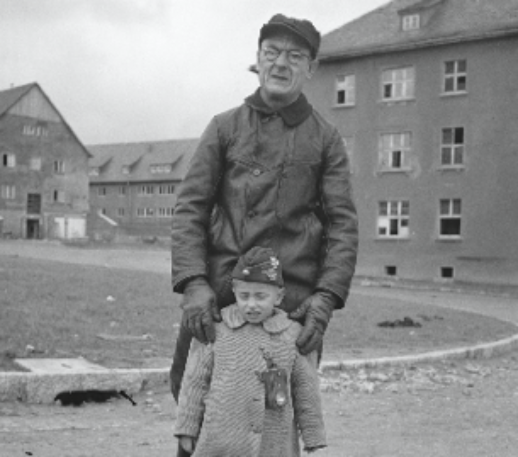 Dieses Foto zeigt einen älteren Mann und ein kleines Kind, die gemeinsam draußen vor einem Gebäude stehen. Die Umgebung wirkt wie eine ehemalige Lager- oder Kasernenanlage – erkennbar an den mehrstöckigen, schlichten Gebäuden im Hintergrund. Der Mann trägt eine einfache, abgenutzte Jacke, Mütze und Handschuhe – seine Kleidung und sein Gesichtsausdruck deuten auf ein hartes Leben hin, möglicherweise als Zwangsarbeiter oder Häftling. Das Kind trägt einen dicken Mantel und eine Mütze mit kleinen Abzeichen oder Ansteckern, die nicht eindeutig zu erkennen sind. Auffällig ist das Schild oder die Marke, die an der Kleidung des Kindes hängt – möglicherweise ein Namensschild oder eine Kennzeichnung, wie sie bei evakuierten oder befreiten Kindern verwendet wurde. Das Bild vermittelt einen sehr menschlichen, berührenden Moment: die Beziehung zwischen dem älteren Mann und dem Kind – vielleicht Großvater und Enkel – steht im Kontrast zu der kargen und beschädigten Umgebung. Es symbolisiert Hoffnung und familiären Zusammenhalt trotz schwerster Umstände.