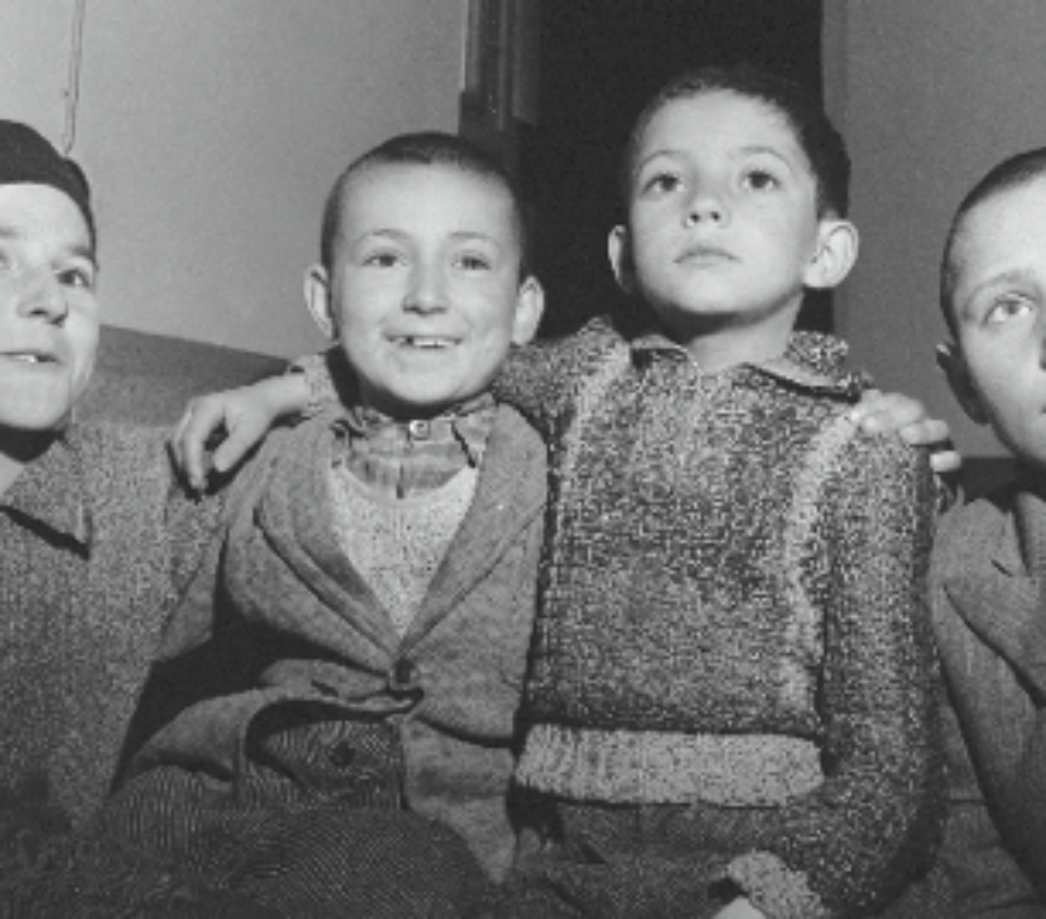 Dieses Bild zeigt vier Jungen, die eng beieinandersitzen und teilweise die Arme umeinander gelegt haben. Sie tragen einfache, aber warme Kleidung – Strickpullover, Jacken, Hosen –, was darauf hinweist, dass sie versorgt wurden, möglicherweise nach der Befreiung eines Lagers oder in einem Auffanglager. Die Gesichtsausdrücke sind gemischt: Einer lacht, ein anderer blickt ernst und konzentriert nach oben. Ihre kurzen Haare deuten auf eine kürzliche Rasur hin, wie sie in Lagern oder bei medizinischer Versorgung üblich war – oft aus hygienischen Gründen. Obwohl die Vergangenheit dieser Kinder vermutlich von Leid geprägt war, vermittelt das Bild durch ihre Nähe zueinander und den Moment des Innehaltens auch ein Gefühl von Hoffnung und Zusammenhalt. Es ist ein eindrucksvolles Zeugnis dafür, wie Kinder selbst unter extremsten Bedingungen Menschlichkeit und Verbindung bewahren können.
