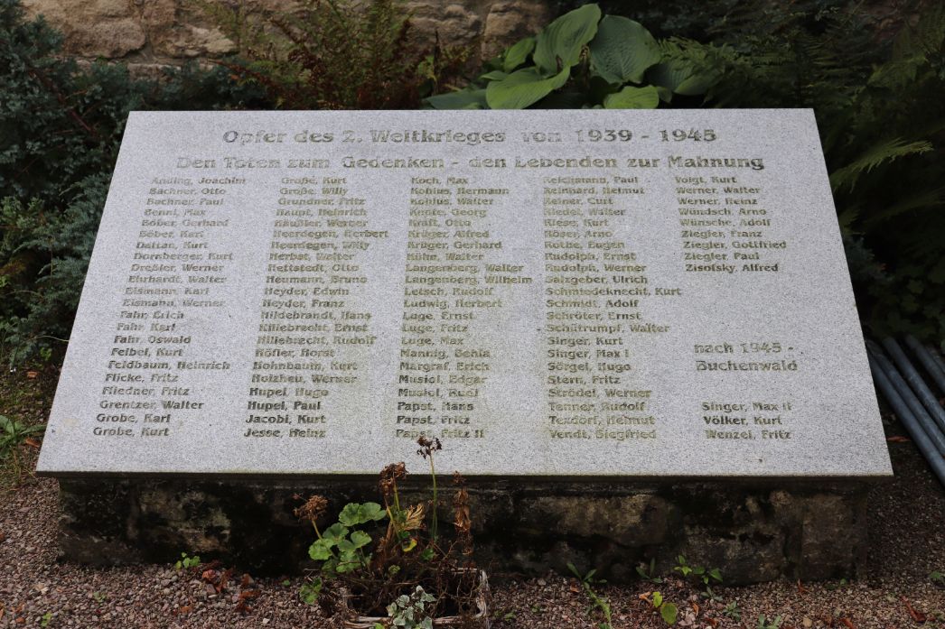 Gedenktafel aus Stein für die „Opfer des 2. Weltkrieges von 1939–1945“. Die Inschrift lautet: „Den Toten zum Gedenken – den Lebenden zur Mahnung“. Auf der Tafel sind zahlreiche Namen von gefallenen oder vermissten Personen alphabetisch eingraviert. In der unteren rechten Ecke wird mit „nach 1945 – Buchenwald“ auf weitere Opfer verwiesen. Die Tafel ist in einem Gartenbereich zwischen Pflanzen aufgestellt.