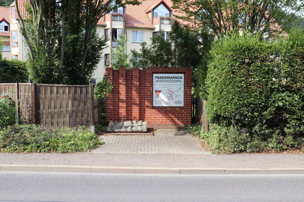 Gedenkstelle am Straßenrand mit roter Backsteinwand und einer Informationstafel zum Todesmarsch von Häftlingen aus dem KZ Buchenwald im April 1945. Die Tafel zeigt eine Karte mit Marschrouten. Davor liegt ein stilisiertes Relief aus Stein, das an menschliche Körper erinnert. Die Gedenkstätte ist von Hecken und Wohnhäusern umgeben.
