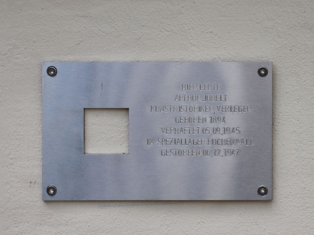Gedenktafel aus Metall an einer Hauswand. In der Mitte befindet sich ein quadratisches, leeres Feld, das möglicherweise auf einen früheren Stolperstein hinweist. Rechts davon ist eingraviert: „Hier lebte Arthur Jubelt Kunsthistoriker, Verleger Geboren 1894 Verhaftet 05.09.1945 im Speziallager Buchenwald Gestorben 06.12.1947“ Die Tafel ist mit vier Schrauben befestigt.