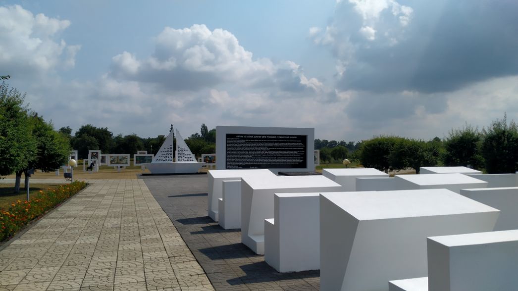 Das Bild zeigt einen Teil der Gedenkstätte Trostenez (auch Maly Trostinec) nahe Minsk in Belarus – einem der zentralen Orte des Holocausts in Osteuropa. Beschreibung: Im Vordergrund stehen mehrere weiße, stilisierte Blöcke, die an Sarkophage oder Gräber erinnern – möglicherweise symbolisieren sie Massengräber oder zerstörte jüdische Gemeinden. Dahinter befindet sich eine dreieckige Stele mit vielen kleinen Aussparungen, die an Namenstafeln oder Lichter erinnern. In der Mitte: Eine große schwarze Gedenktafel mit einem längeren Text auf Russisch (vermutlich auch Belarussisch und/oder Englisch) – wahrscheinlich eine Erklärung der historischen Ereignisse und ein Gedenken an die Opfer. Im Hintergrund auf dem Rasen: Weitere Tafeln mit Bildern und Texten – offenbar eine Ausstellung zur Geschichte oder zur Erinnerungskultur.