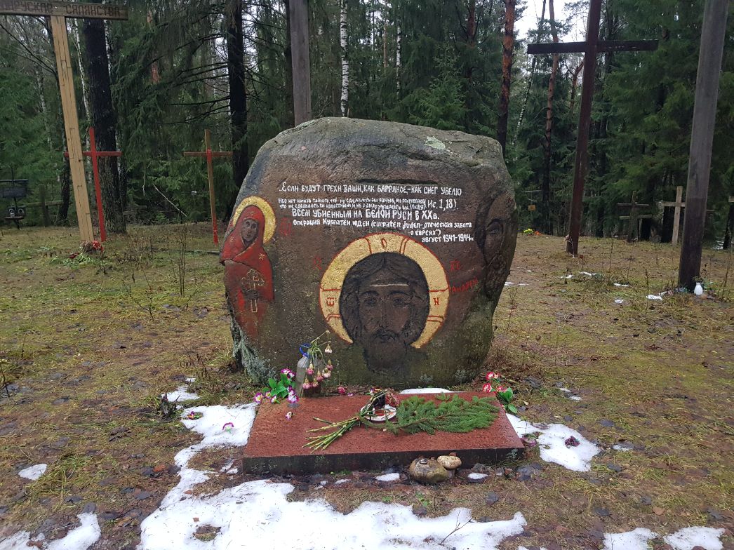 Das Bild zeigt ein Gedenkstein-Ensemble im Wald – vermutlich eine Gedenkstätte für Opfer nationalsozialistischer Gewaltverbrechen oder Kriegsopfer in Belarus. Beschreibung des Bildes: Zentral steht ein großer Naturstein, in den christlich-orthodoxe Ikonen (Christus Pantokrator, Muttergottes) und ein Text eingemeißelt und farbig bemalt sind. Der Text ist in russischer Sprache gehalten und verweist auf ein Massaker an der belarussischen Zivilbevölkerung im Zuge der Operation "Kottbus" – einer sog. „Bandenbekämpfungsaktion“ der deutschen Wehrmacht und SS im Jahr 1943. Rundherum sind orthodoxe Kreuze aus Holz zu sehen, die wie Grabkreuze oder Gedenkkreuze aufgestellt sind. Vor dem Gedenkstein liegen Blumen, Kerzen, kleine Andenken und Tannenzweige – Zeichen individuellen Gedenkens. Im Hintergrund: dichter Nadelwald, stellenweise noch mit Schnee bedeckt – deutet auf Winter oder frühen Frühling hin. Textauszug auf dem Stein (sinngemäß übersetzt): „Und wenn eure Sünden rot wie Scharlach sind, werden sie weiß wie Schnee … (Jes 1,18). Alle Bewohner des Dorfes Belye Berega wurden hier lebendig verbrannt während der deutschen Strafaktion 'Kottbus' (Mai–Juni 1943).“