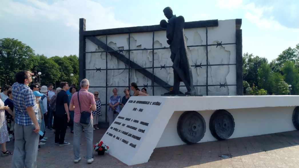 Das Bild zeigt ein Holocaust-Mahnmal in Maly Trostinez, nahe Minsk in Belarus, das an die Opfer des nationalsozialistischen Vernichtungslagers erinnert. Es handelt sich um eines der wichtigsten Denkmäler auf dem Gelände des ehemaligen Lagers. Beschreibung des Denkmals: Im Vordergrund ist eine Figur eines trauernden Mannes, der ein totes Kind im Arm hält – ein starkes Symbol für Leid, Verlust und Menschlichkeit im Angesicht der Vernichtung. Die Figur steht auf einem Sockel, der wie ein Waggon oder Transportmittel gestaltet ist – ein Verweis auf die Deportationen. Die Wand im Hintergrund zeigt schemenhafte Menschenfiguren hinter angedeutetem Stacheldraht, was die Bedingungen im Lager symbolisiert. Auf dem Sockel ist ein Text in Russisch zu lesen: „Allen, die nicht aus dem Krieg zurückkehrten. 1941–1945.“ Darunter eine Widmung an ermordete Zivilisten, Kinder, ältere Menschen und ganze Familien, die hier von den Nationalsozialisten ermordet wurden.