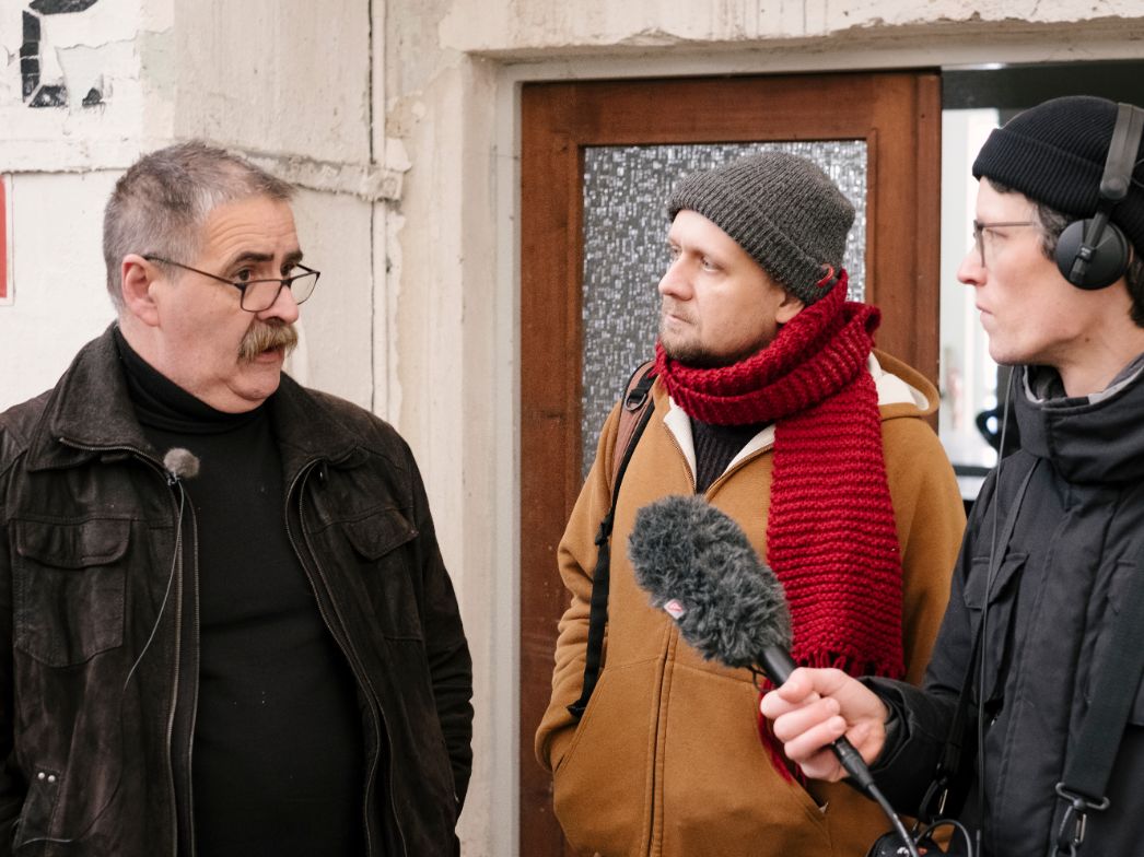 Drei Männer stehen in einem Innenraum vor einer Tür. Der Mann links trägt eine dunkle Jacke, Brille und ein Mikrofon am Revers, er spricht. Der Mann in der Mitte trägt eine graue Mütze, einen roten Strickschal und eine braune Jacke, er hört aufmerksam zu. Der Mann rechts trägt Kopfhörer und hält ein Mikrofon mit Windschutz in Richtung des Sprechers.