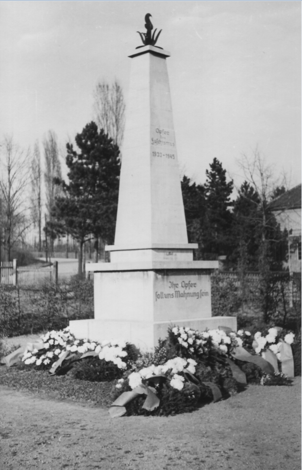 Eingangsbereich des Ehrenfriedhofs Nordhausen mit moderner Gedenkarchitektur – im Vordergrund eine große, blaue Stele mit der Aufschrift ‚Ehrenfriedhof Nordhausen‘, im Hintergrund ein heller Torbau mit Informationstafeln, umgeben von Rasenflächen und Bäumen.