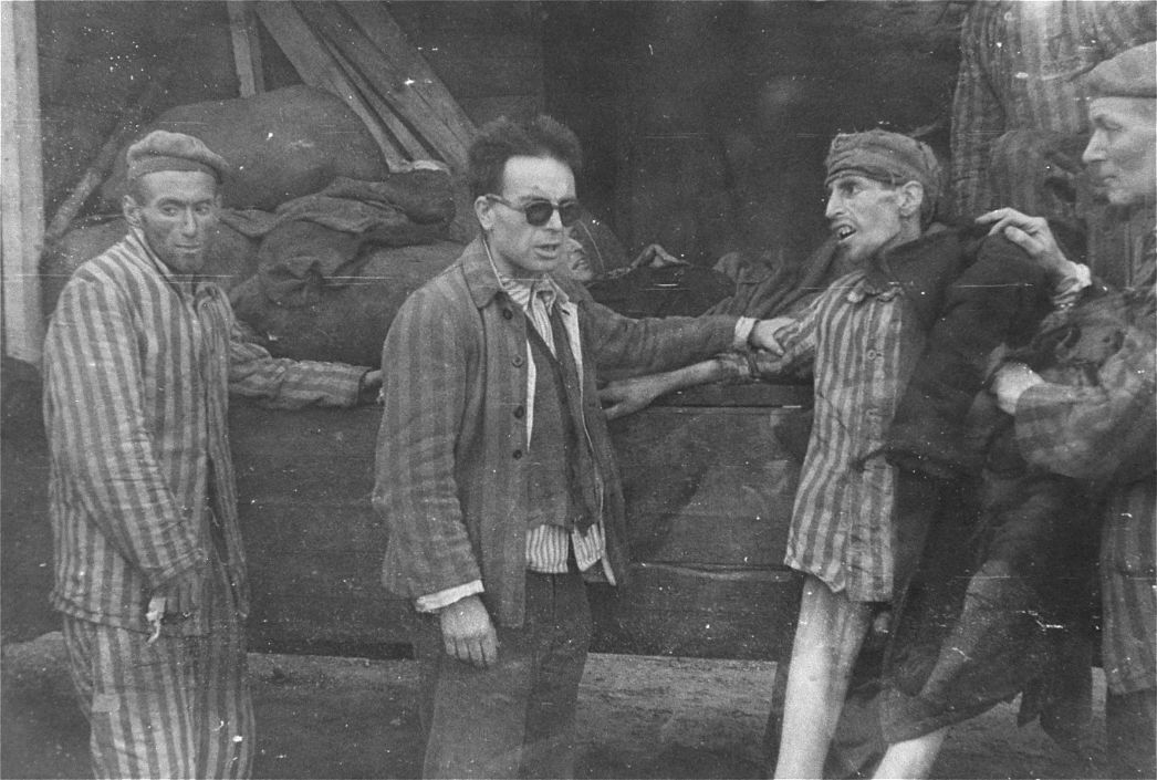 Schwarz-weiß-Fotografie von überlebenden Häftlingen des Konzentrationslagers Buchenwald nach der Befreiung im April 1945. Im Zentrum steht ein Mann mit Sonnenbrille in gestreifter Häftlingskleidung, der einen schwer abgemagerten Mithäftling stützt. Weitere Männer in Häftlingsuniformen sind im Bild zu sehen. Im Hintergrund liegen ausgemergelte Körper auf einem offenen Lastwagen. Die Szene vermittelt eindrücklich das Ausmaß des physischen und seelischen Leids der Lagerinsassen.