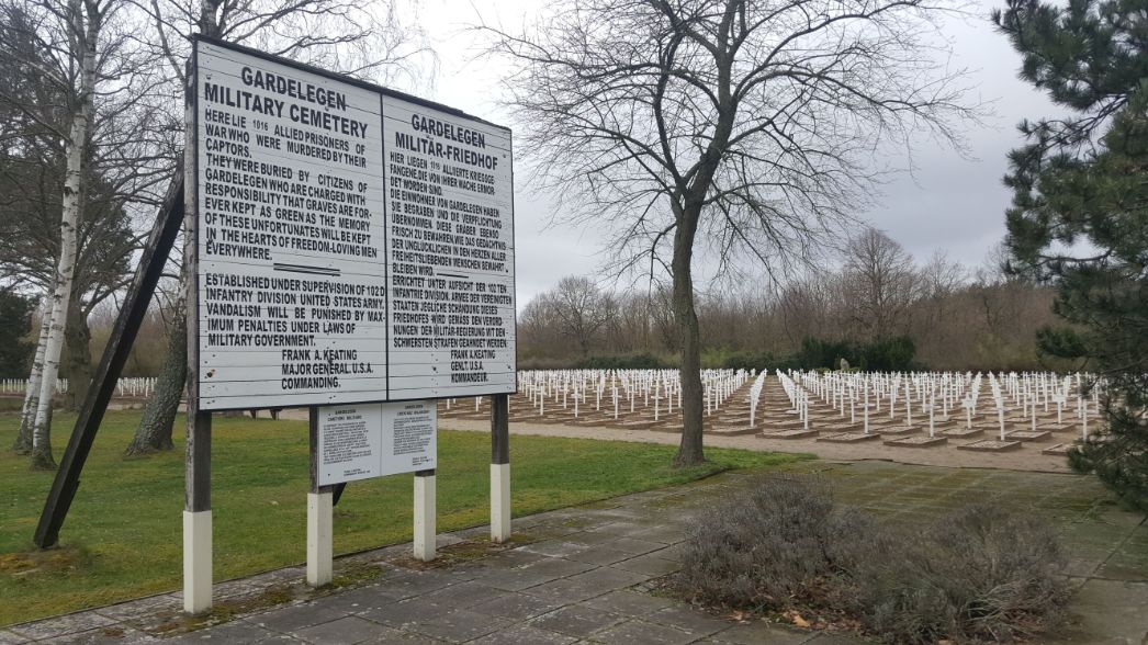 Ein Schild kennzeichnet den Militärfriedhof von Gardelegen. Dahinter erstreckt sich ein großes Feld mit vielen gleichförmigen weißen Kreuzen als Grabzeichen.