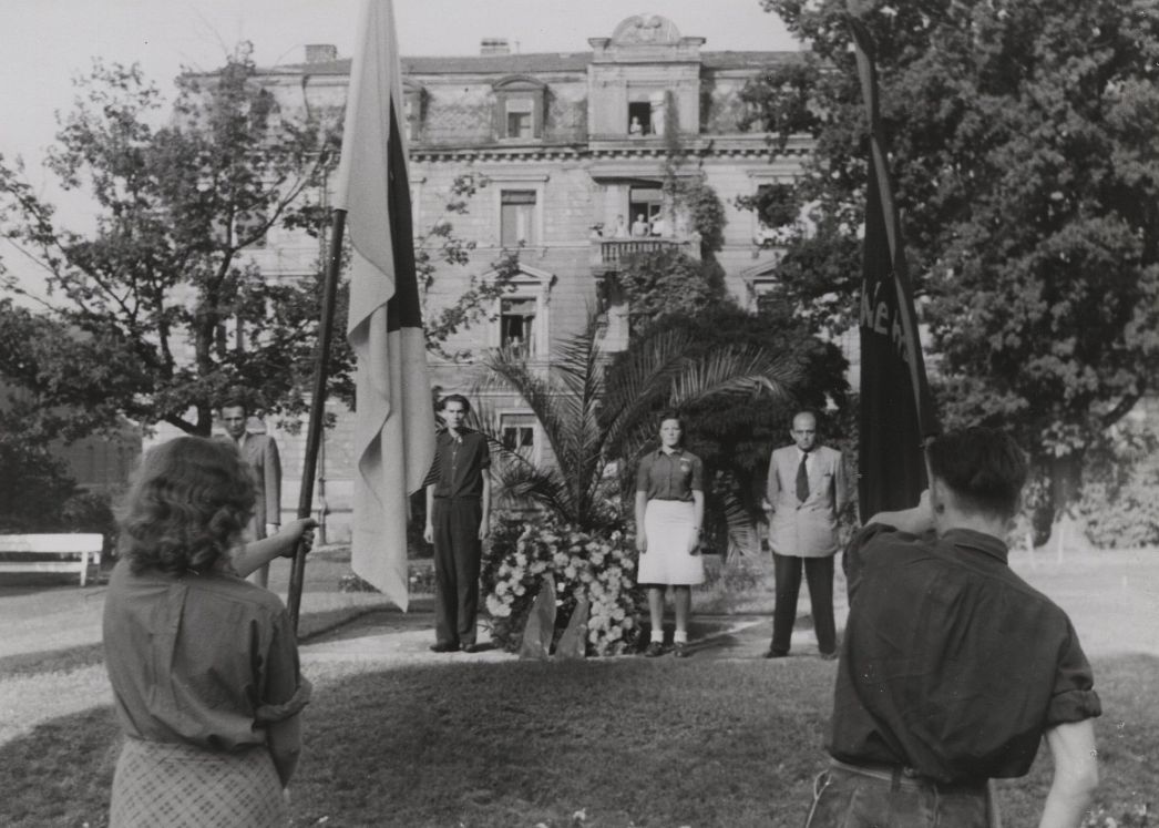 Historische Schwarzweißaufnahme einer Gedenkzeremonie im Freien. Im Vordergrund stehen zwei Personen mit Fahnen, die auf ein Blumenarrangement in der Mitte gerichtet sind. Dahinter haben sich mehrere Männer und eine Frau in einer Reihe aufgestellt und blicken nach vorn. Im Zentrum liegt ein großer Kranz mit Blumen. Im Hintergrund erhebt sich ein mehrstöckiges Gebäude mit Balkon und geöffneten Fenstern, teilweise von Bäumen und Pflanzen umgeben.