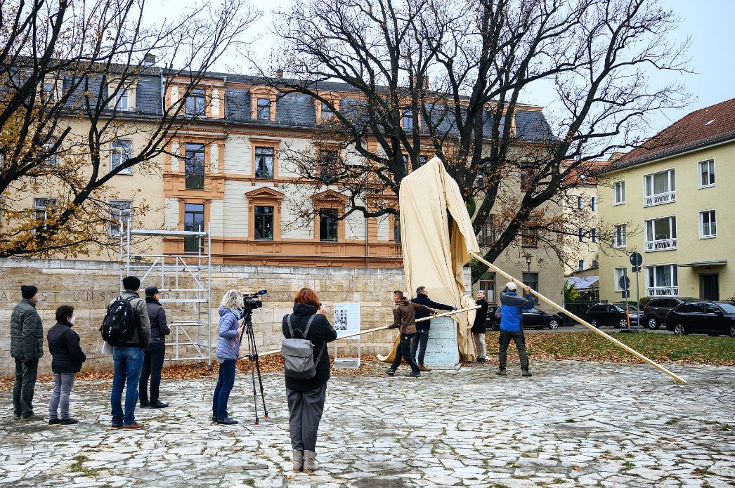Mehrere Menschen stehen auf einem gepflasterten Platz und beobachten, wie ein verhülltes Denkmal enthüllt wird. Einige Personen ziehen mit langen Holzstangen eine große helle Stoffhülle von einer Statue oder Stele herunter. Eine Person filmt die Szene mit einer Kamera auf einem Stativ. Im Hintergrund stehen Bäume ohne Laub sowie mehrstöckige Wohn- und Verwaltungsgebäude.