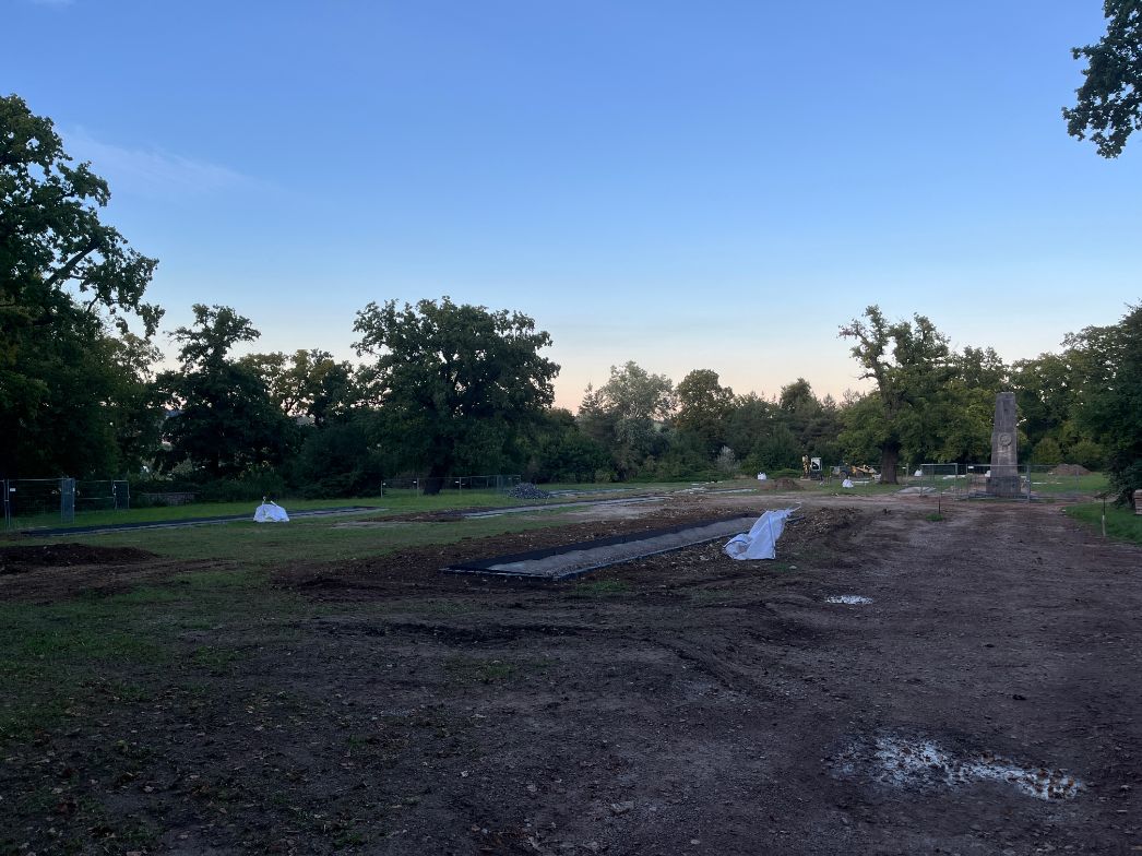 Weitläufiges Außengelände, das wie eine Baustelle oder ein neu gestalteter Friedhofsbereich wirkt. Der Boden ist teilweise aufgerissen, mit Erdflächen, Schotter und angelegten rechteckigen Gräben oder Fundamenten. Vereinzelt liegen mit Planen abgedeckte Objekte. Im Hintergrund stehen Bäume und Sträucher, rechts ist ein aufragendes Denkmal oder Obelisk zu sehen, umgeben von Bauzäunen und Arbeitsgeräten. Der Himmel ist klar und weit.