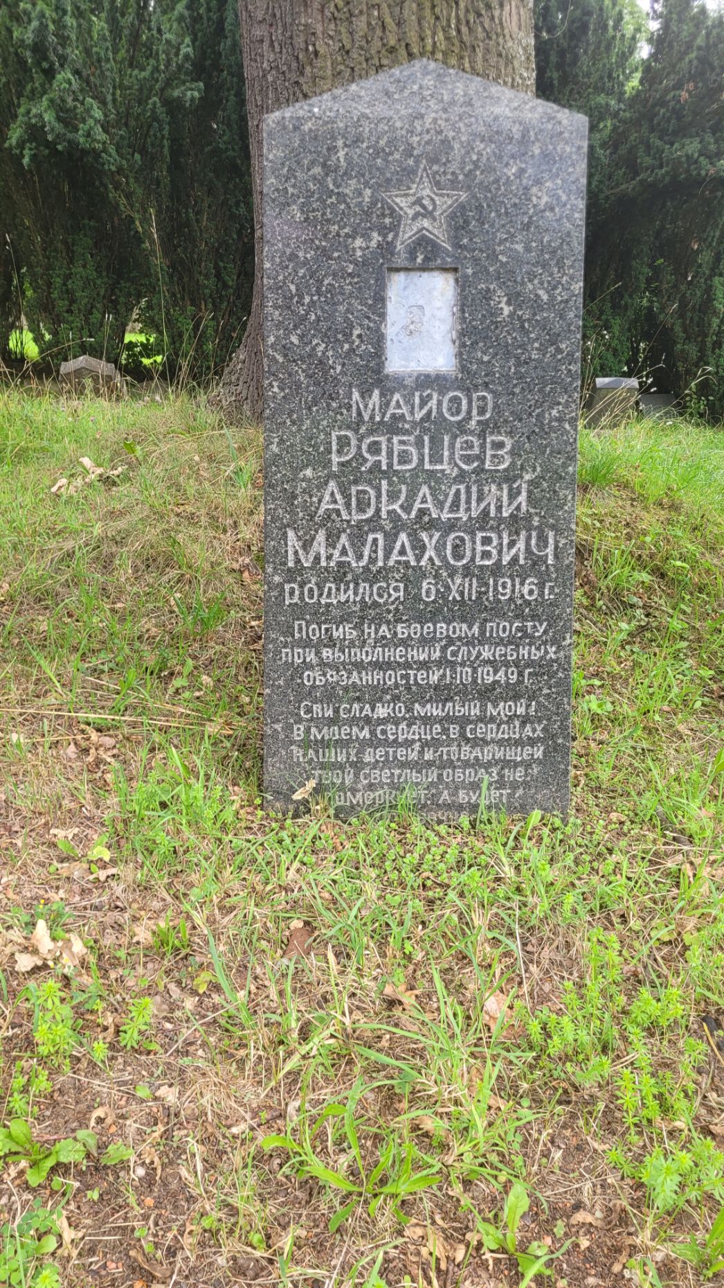 Aufrechter Grabstein aus dunklem Stein auf einer Rasenfläche, im Hintergrund Bäume. Oben ist ein eingravierter Stern mit Hammer-und-Sichel-Symbol zu sehen, darunter eine kleine rechteckige Vertiefung für ein Porträt. Die Inschrift ist in kyrillischer Schrift gehalten und nennt einen Offizier (Major) mit Namen sowie Geburts- und Todesangaben.