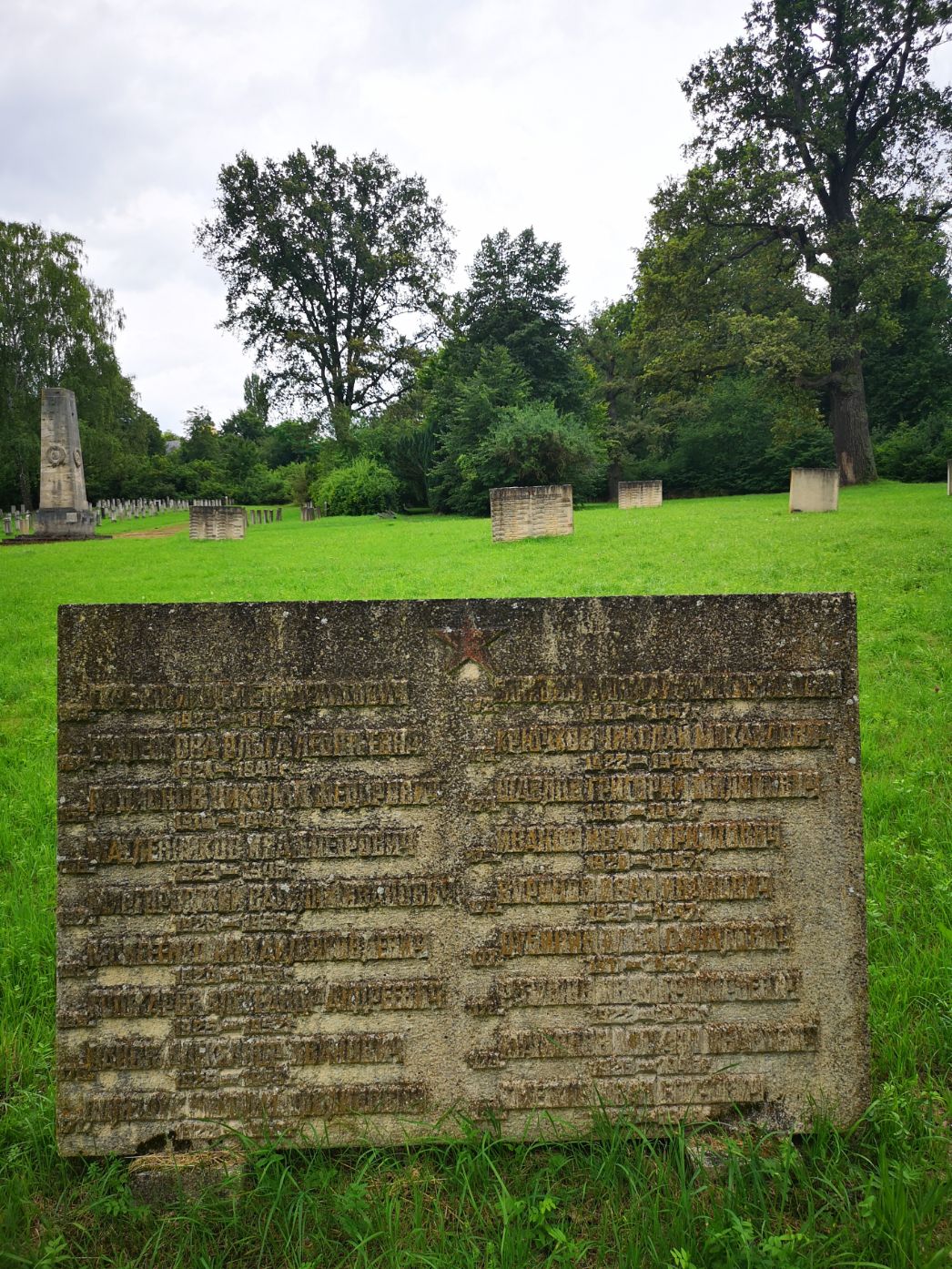 Großer steinerner Gedenkblock mit verwitterter Oberfläche und kaum noch lesbarer kyrillischer Inschrift im Vordergrund einer weitläufigen Rasenfläche. Dahinter stehen mehrere weitere niedrige Steinblöcke in regelmäßigen Abständen. Links ist ein höheres Denkmal oder Obelisk zu sehen, umgeben von zahlreichen kleinen Grabzeichen. Die Umgebung wirkt parkartig mit großen Bäumen und dichtem Grün im Hintergrund unter einem bewölkten Himmel.