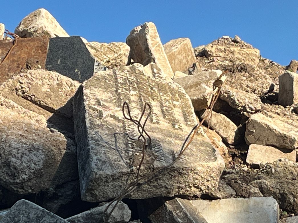 Nahaufnahme eines Haufens aus zerbrochenen Steinplatten und Grabsteinen unter blauem Himmel. Die Steine liegen unregelmäßig übereinander, teils mit sichtbaren Resten von Inschriften. Einige sind gebrochen oder stark verwittert. Zwischen den Steinen ragen rostige Drahtstücke hervor.