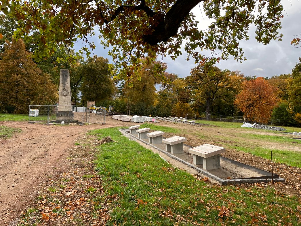 Weitläufiger Friedhofsbereich in einer Parklandschaft im Herbst. Im Vordergrund verläuft eine neu angelegte Reihe von niedrigen, rechteckigen Grab- oder Gedenksteinen entlang eines schmalen, eingefassten Streifens. Links steht ein höheres Denkmal oder Obelisk, der von einem Bauzaun umgeben ist. Im Hintergrund sind Bauarbeiten im Gange, mit Maschinen, Materialien und weiteren neu gesetzten Steinelementen. Große Bäume mit herbstlich gefärbtem Laub rahmen die Szene unter einem bewölkten Himmel.