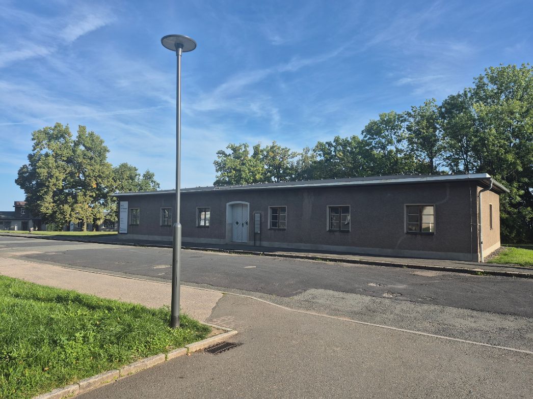 Langes, eingeschossiges Gebäude mit schlichter Fassade und flachem Dach entlang einer asphaltierten Straße. In der Mitte befindet sich ein heller Eingang mit Doppeltür, daneben mehrere kleine Fenster. Vor dem Gebäude steht eine hohe Straßenlaterne. Die Umgebung wirkt ruhig, mit Rasenflächen und Bäumen im Hintergrund unter blauem Himmel.