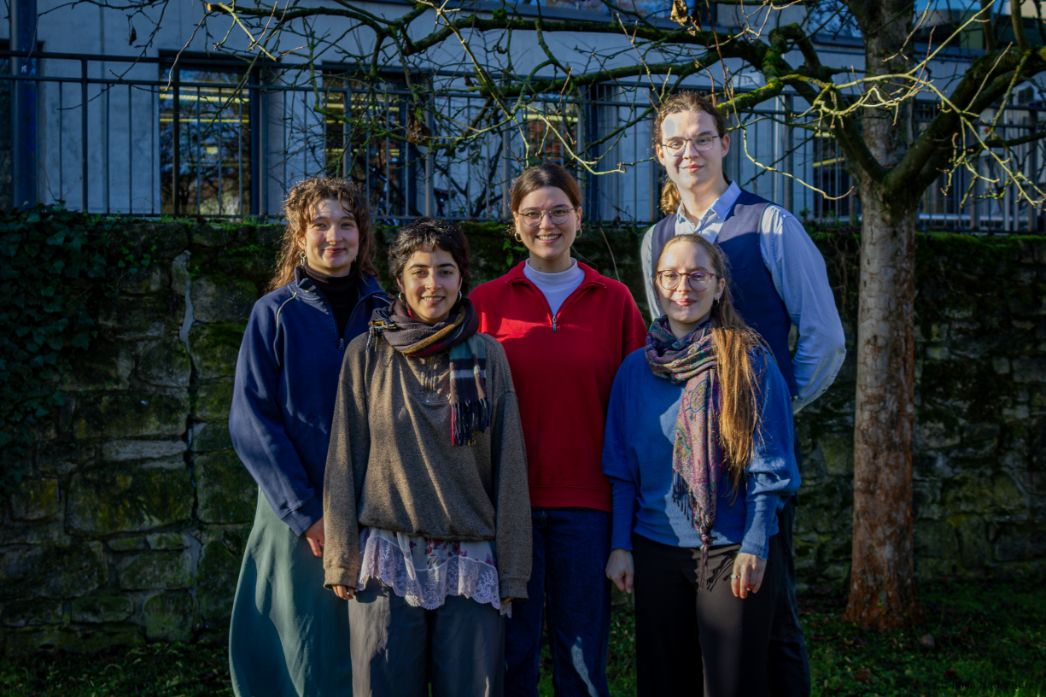 Fünf junge Erwachsene stehen nebeneinander im Freien und blicken in die Kamera. Sie tragen unterschiedliche Alltagskleidung, darunter Pullover, Schal und Hemd, und wirken entspannt und freundlich. Im Hintergrund befinden sich eine niedrige Steinmauer, ein Gebäude mit Fenstern sowie ein Baum mit kahlen Ästen. Die Szene ist von Sonnenlicht geprägt.