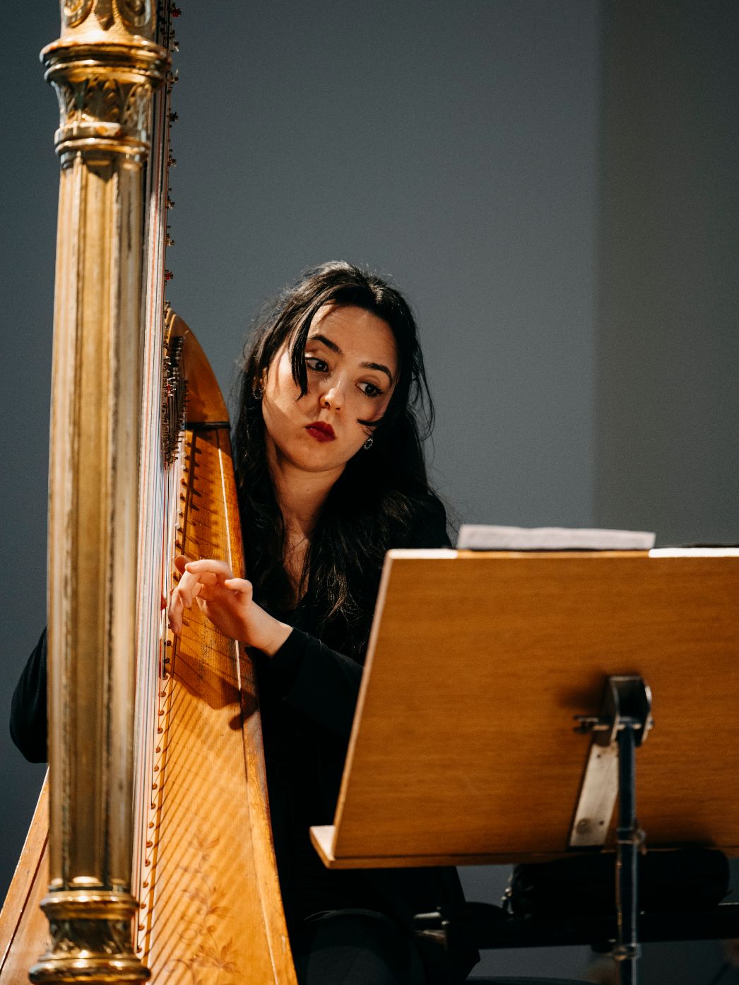 Eine junge Musikerin sitzt konzentriert an einer Harfe und liest gleichzeitig von einem Notenständer. Ihre Haltung und ihr Blick wirken fokussiert und ruhig, während sie die Saiten zupft. Die warme Holzfarbe des Instruments hebt sich vom schlichten Hintergrund ab und lenkt den Blick auf die feine, präzise Bewegung ihrer Hände.