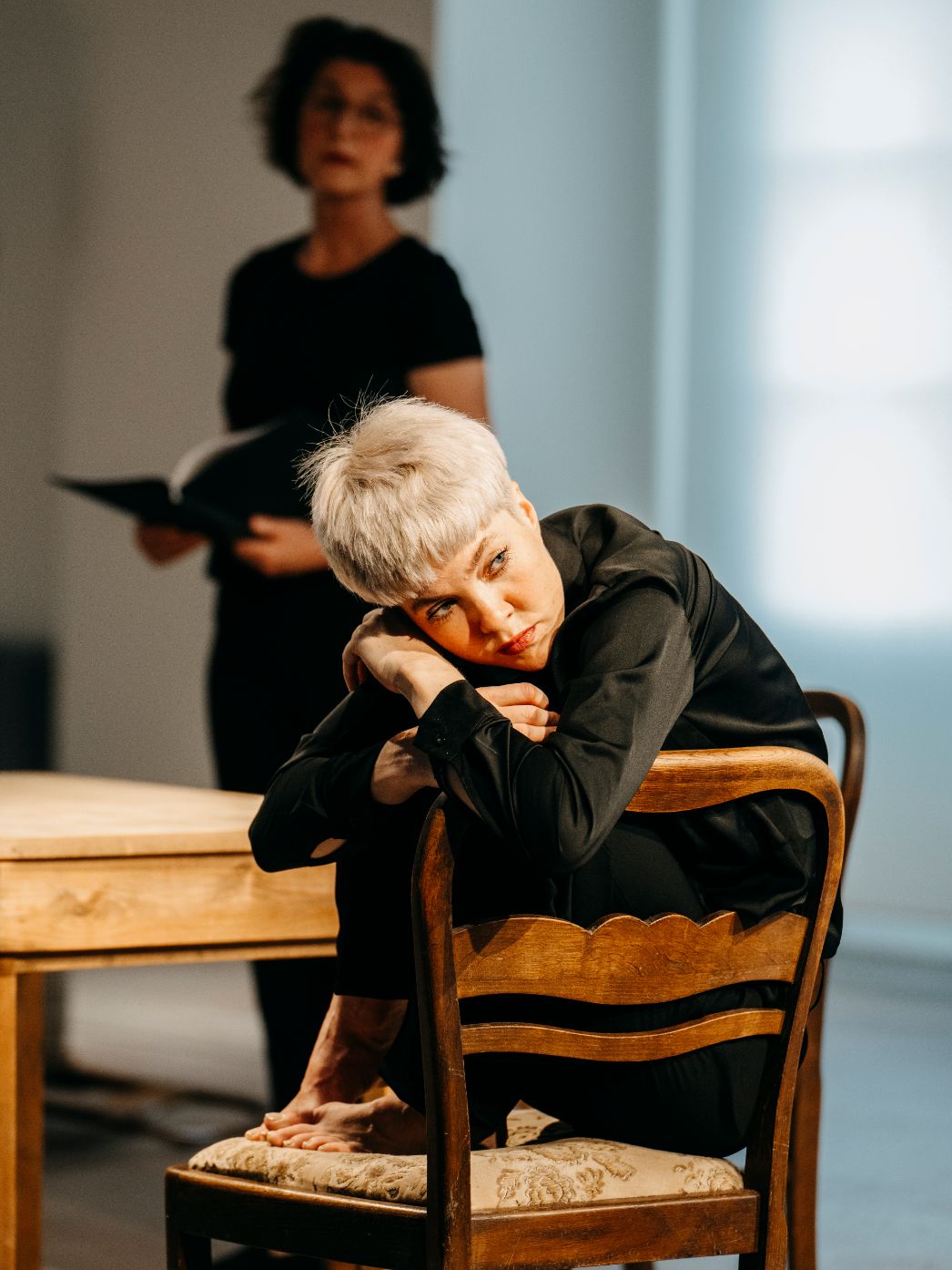 Die Tänzerin sitzt eingekugelt und mit ernstem Blick auf einem Holzstuhl. Sie hat die Arme um sich geschränkt.