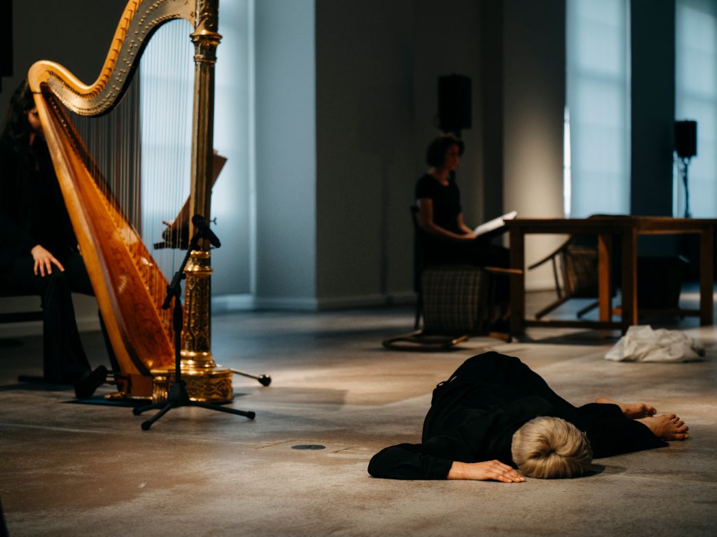 Im Vordergrund liegt eine Person regungslos auf dem Boden, während im Hintergrund eine Harfe gespielt wird und eine weitere Person an einem Tisch sitzt. Die Szene wirkt wie eine inszenierte Performance: Musik, Bewegung und Stillstand stehen in einem spannungsvollen Kontrast. Das gedämpfte Licht und der weite Raum verstärken die ruhige, zugleich nachdenkliche Atmosphäre.