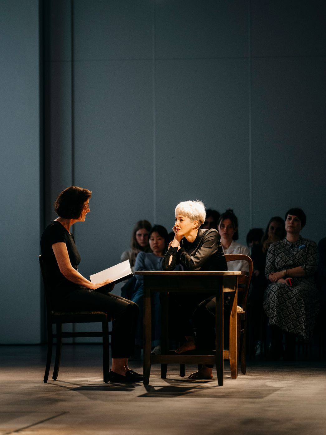 Zwei Frauen sitzen sich an einem Tisch gegenüber und führen ein konzentriertes Gespräch vor Publikum. Die reduzierte Inszenierung und das gerichtete Licht lenken den Fokus auf den Dialog und die Auseinandersetzung mit dem Thema.