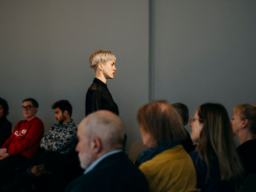 Eine Person mit kurzem, hellem Haar steht aufrecht und ruhig zwischen sitzenden Zuschauer*innen. Ihr seitliches Profil wirkt konzentriert und gesammelt, während das Publikum um sie herum beobachtet. Die Szene hat etwas Performatives: Die klare Haltung der stehenden Figur setzt einen bewussten Kontrast zur sitzenden Menge und lenkt die Aufmerksamkeit auf ihre Präsenz im Raum.